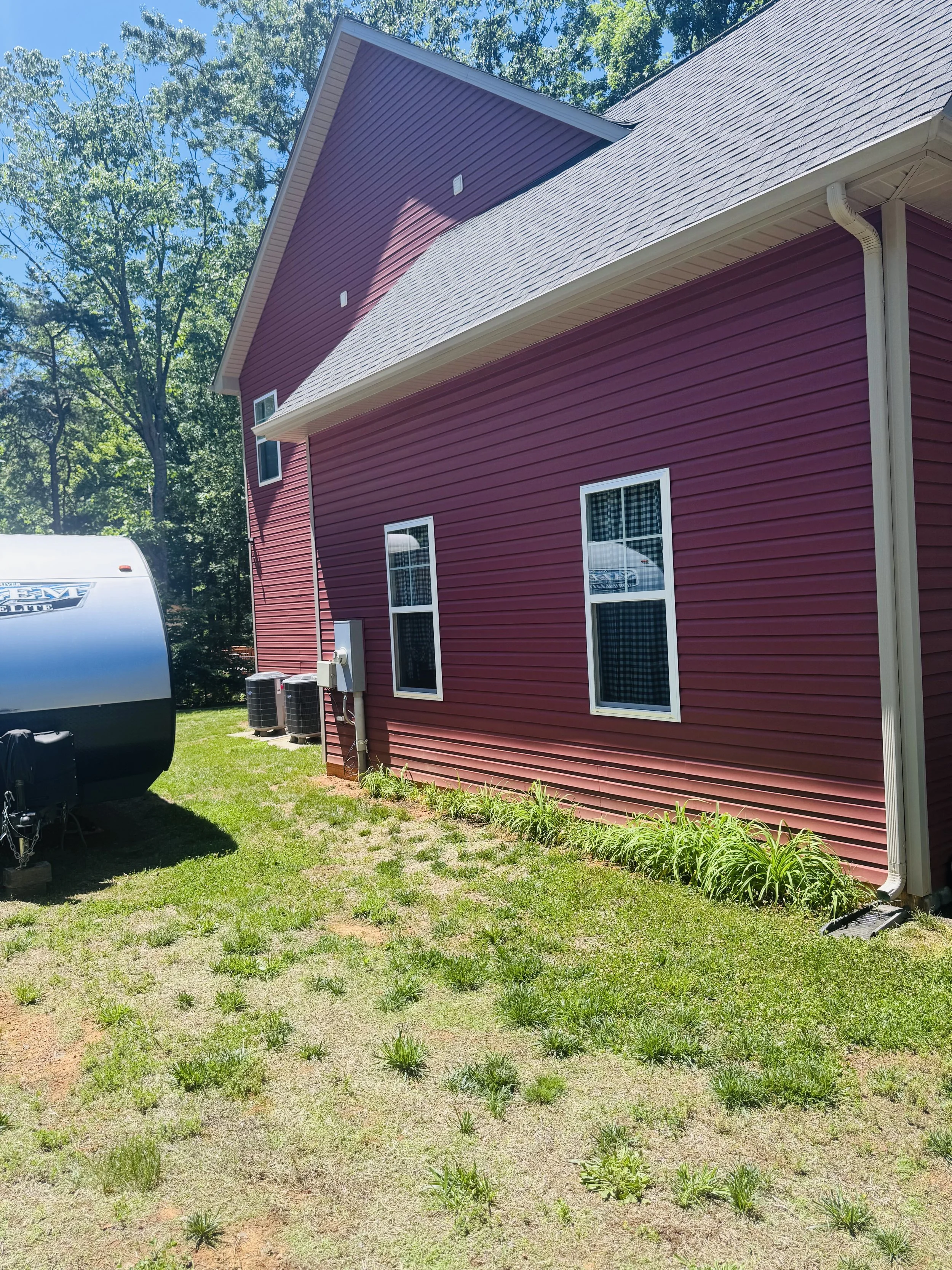 The side of a red house with white trim, three windows, and a beige gutter. Two air conditioning units and a water heater are outside, along with a patch of grass and small plants. In the background, tall green trees and a clear blue sky are visible.