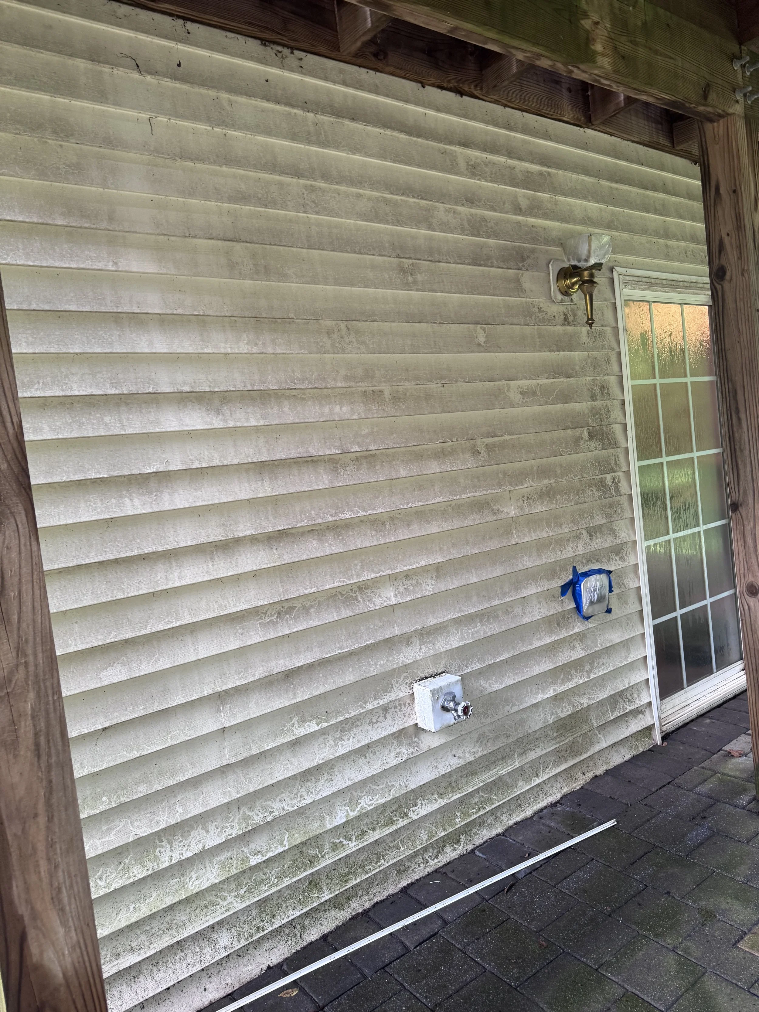 Back porch of a house with dirty beige siding, a window, an outdoor electrical outlet, and a water spigot. The porch is framed with wooden posts and has a brick paver floor.