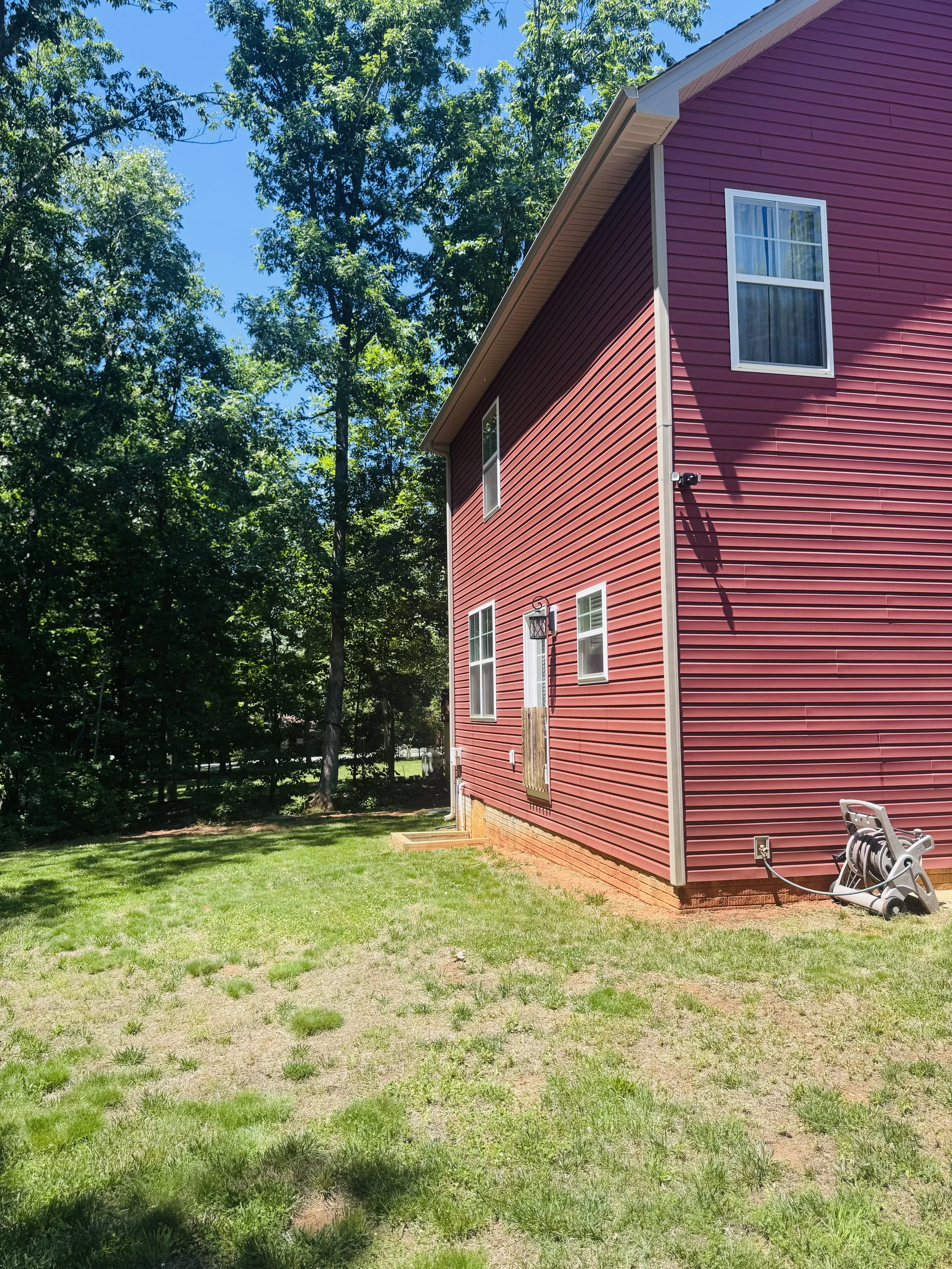 Side view of a red house with white trim, four visible windows, and a lawn with patches of grass and dirt. Large trees are in the background under a clear blue sky.