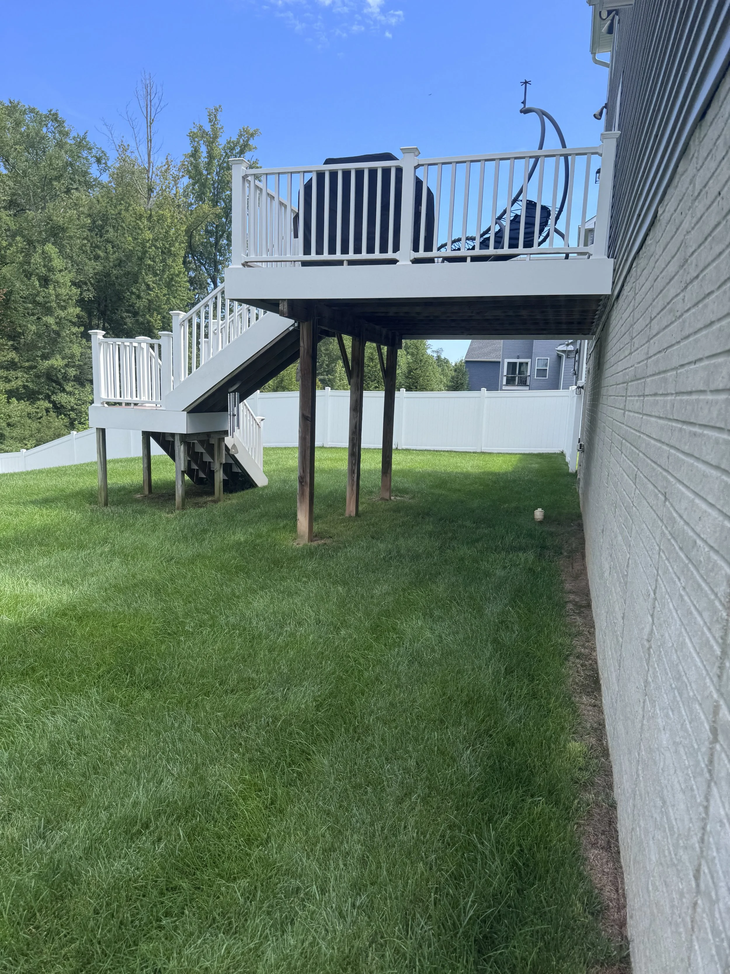 Backyard with a wooden deck and stairs attached to a brick house, green grass, white fence, trees, and a blue sky.