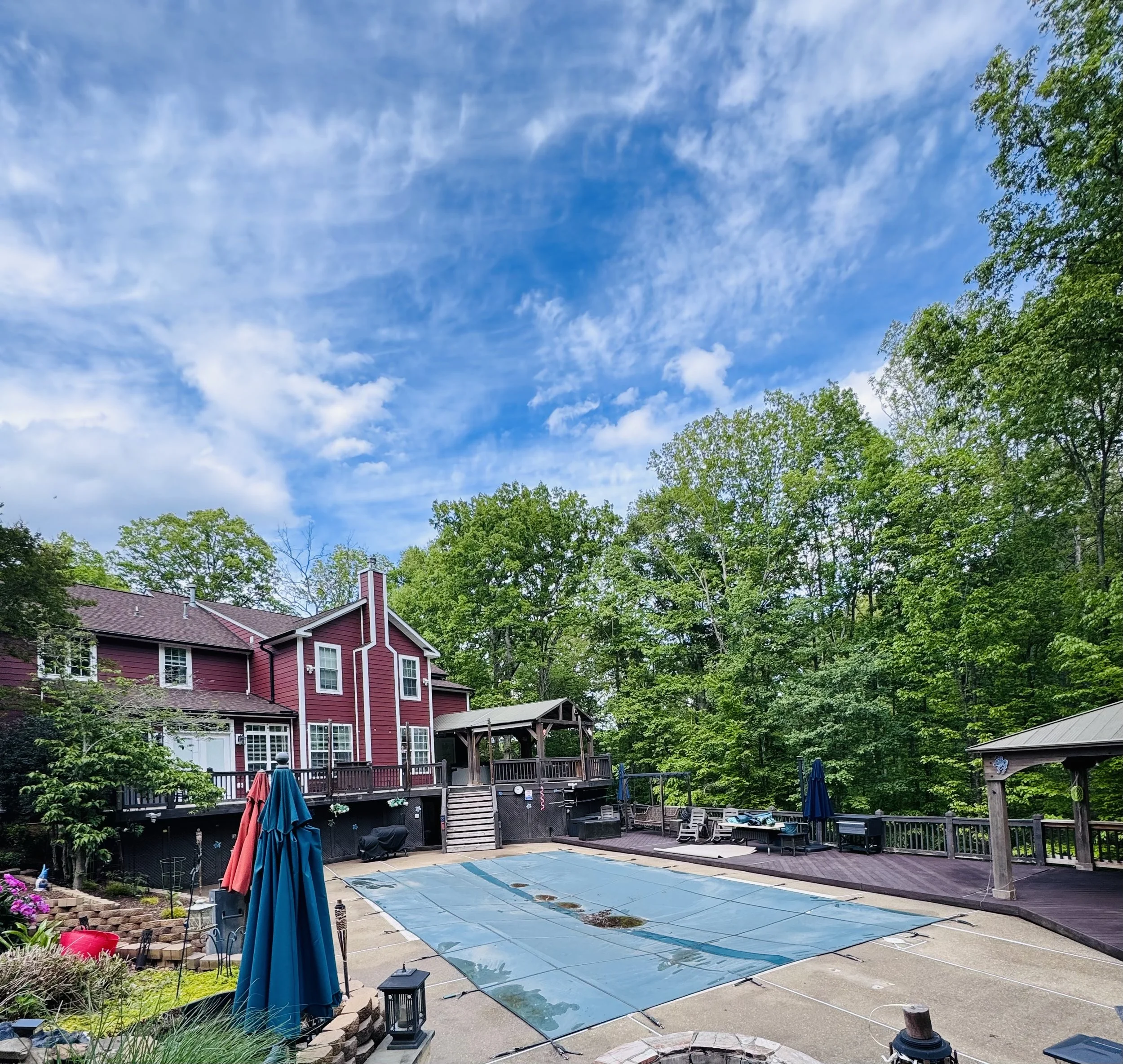 A backyard with a pool covered by a blue safety cover, surrounded by patio furniture, umbrellas, and landscaping, with a red house and tall trees under a partly cloudy sky.
