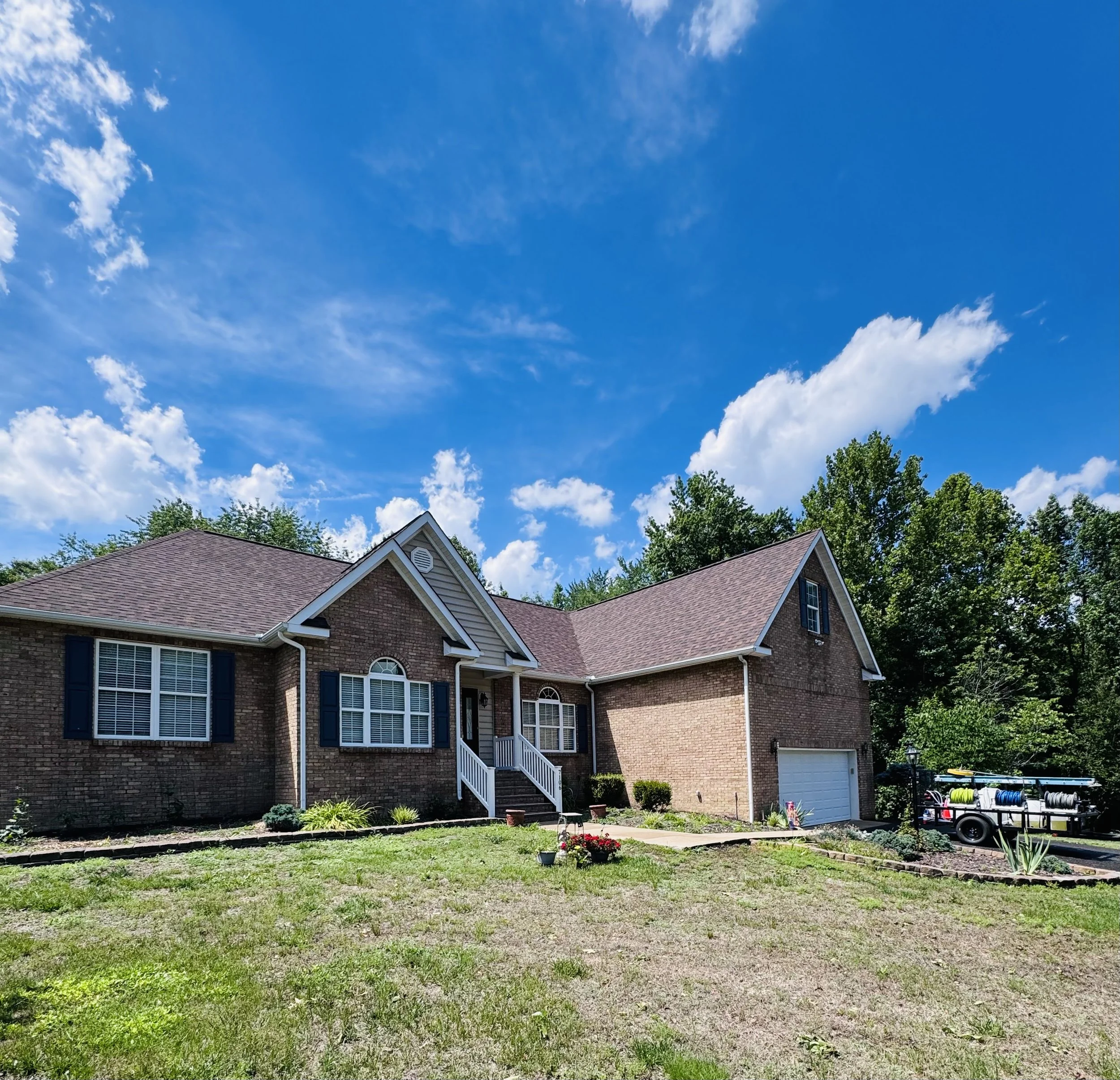View of a large brick house with a brown shingle roof, white trim, and multiple windows. The house is surrounded by green trees and a backyard with a grass, garden beds, and gardening tools. The sky is blue with some scattered clouds.