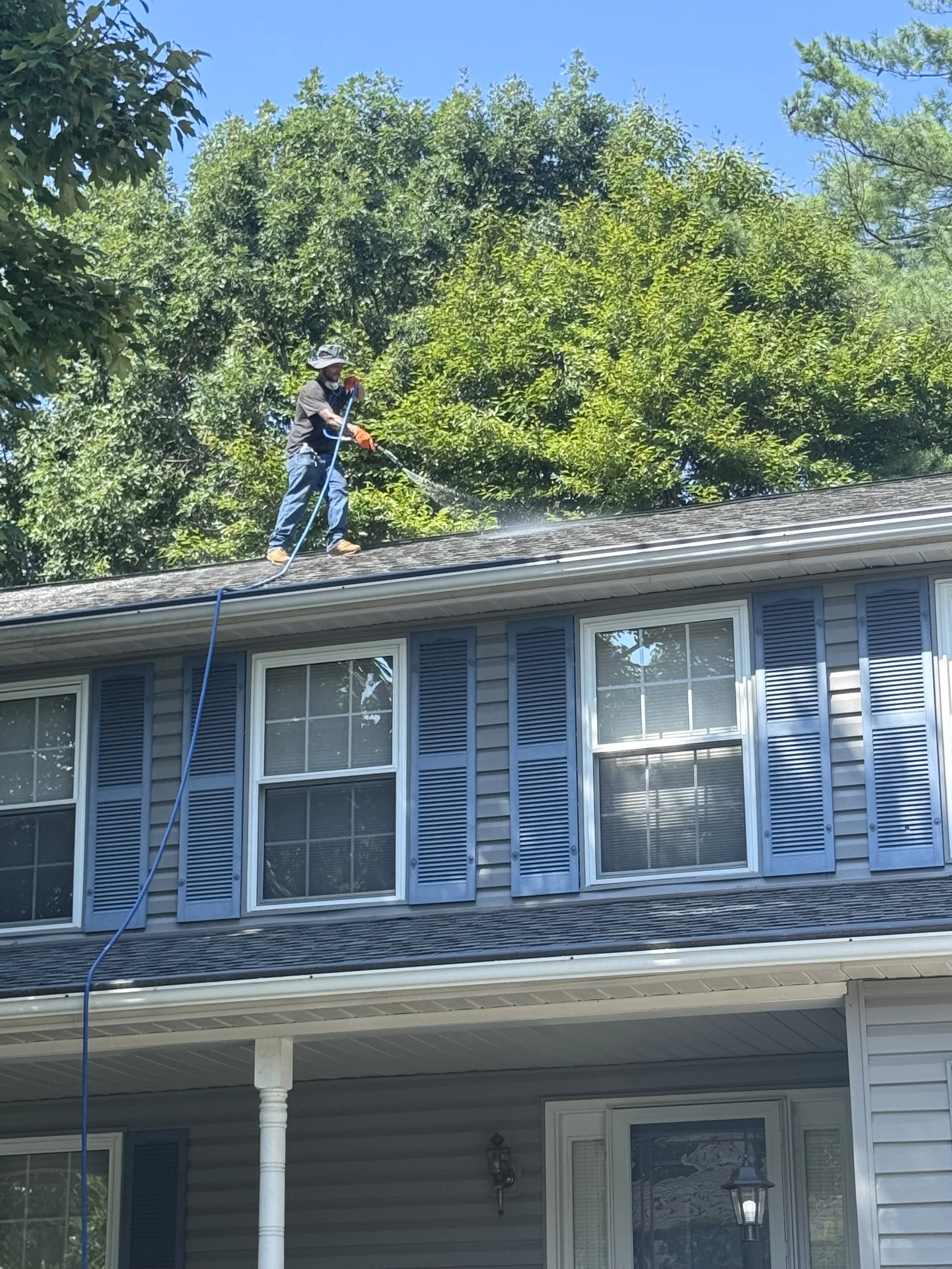 A man wearing a hat, gloves, and casual clothing is on the roof of a house, using a hose to spray water on the roof. The house is a two-story building with blue siding and multiple windows. The scene is outdoors with trees in the background and a cle