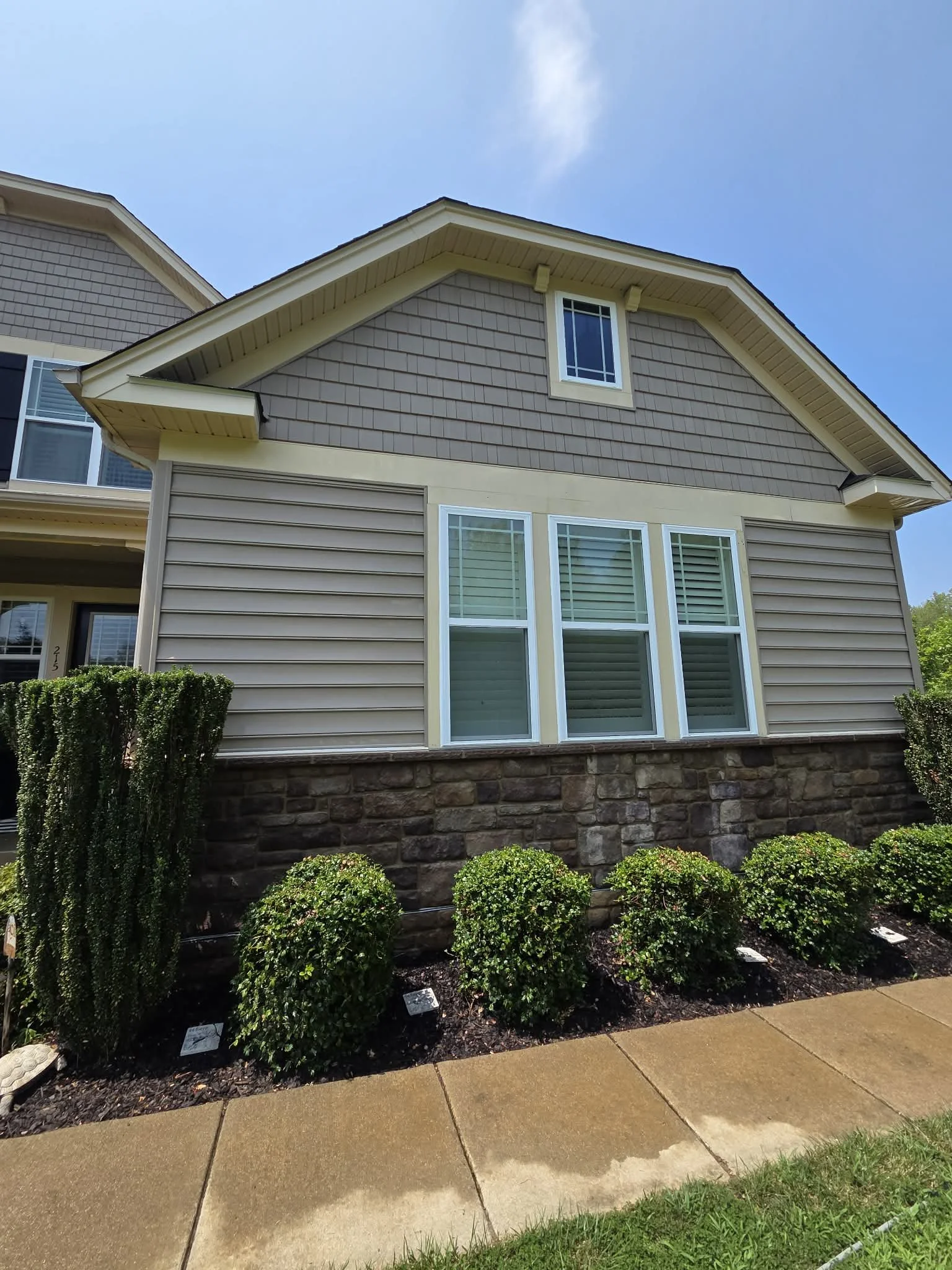 Front view of a modern house with beige siding, a stone foundation, and tall windows, with manicured bushes and a sidewalk in front, under a clear blue sky.