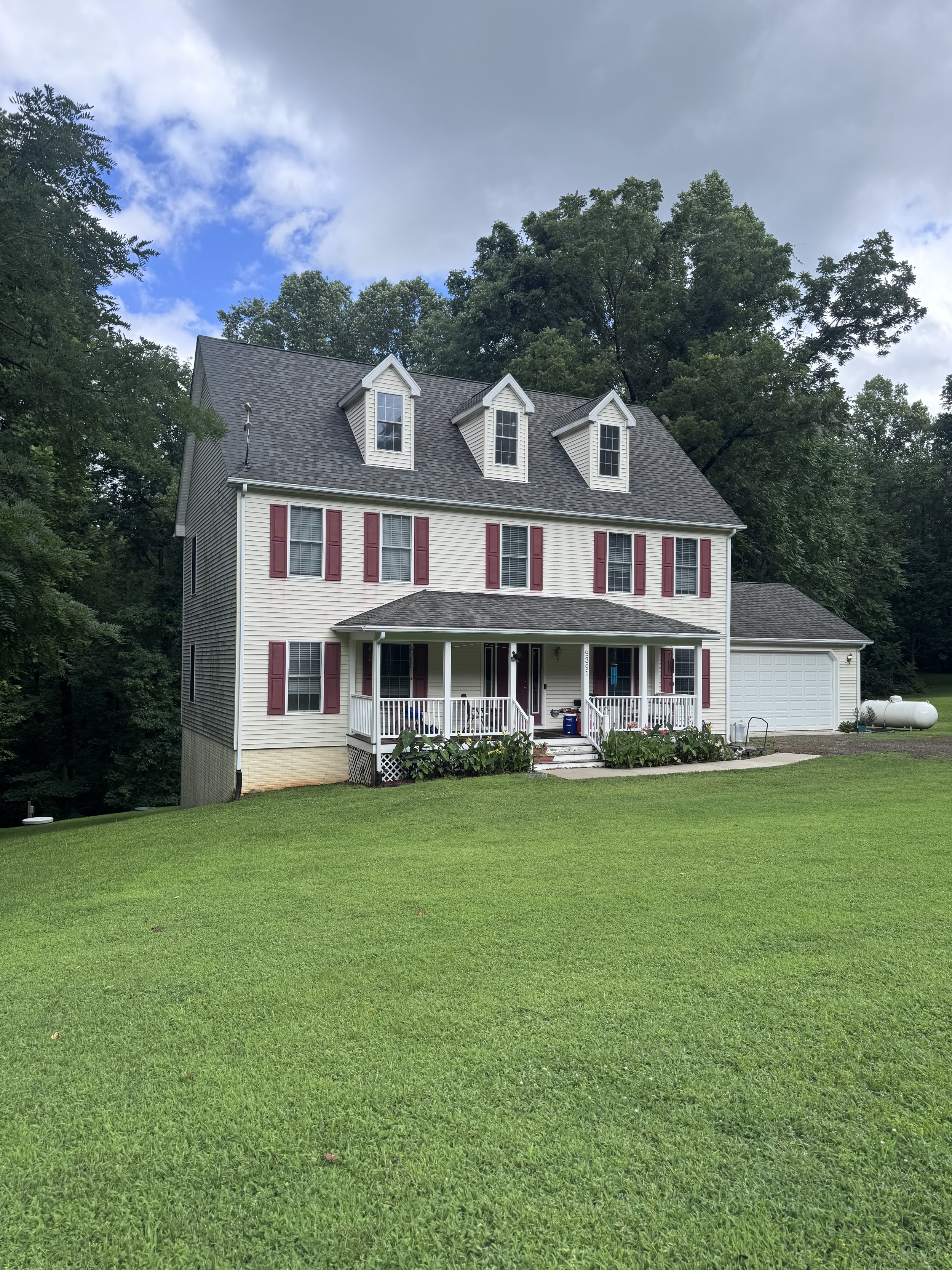 A white three-story house with red shutters, a front porch, and a detached garage, set in a lush green yard with trees in the background under a partly cloudy sky.