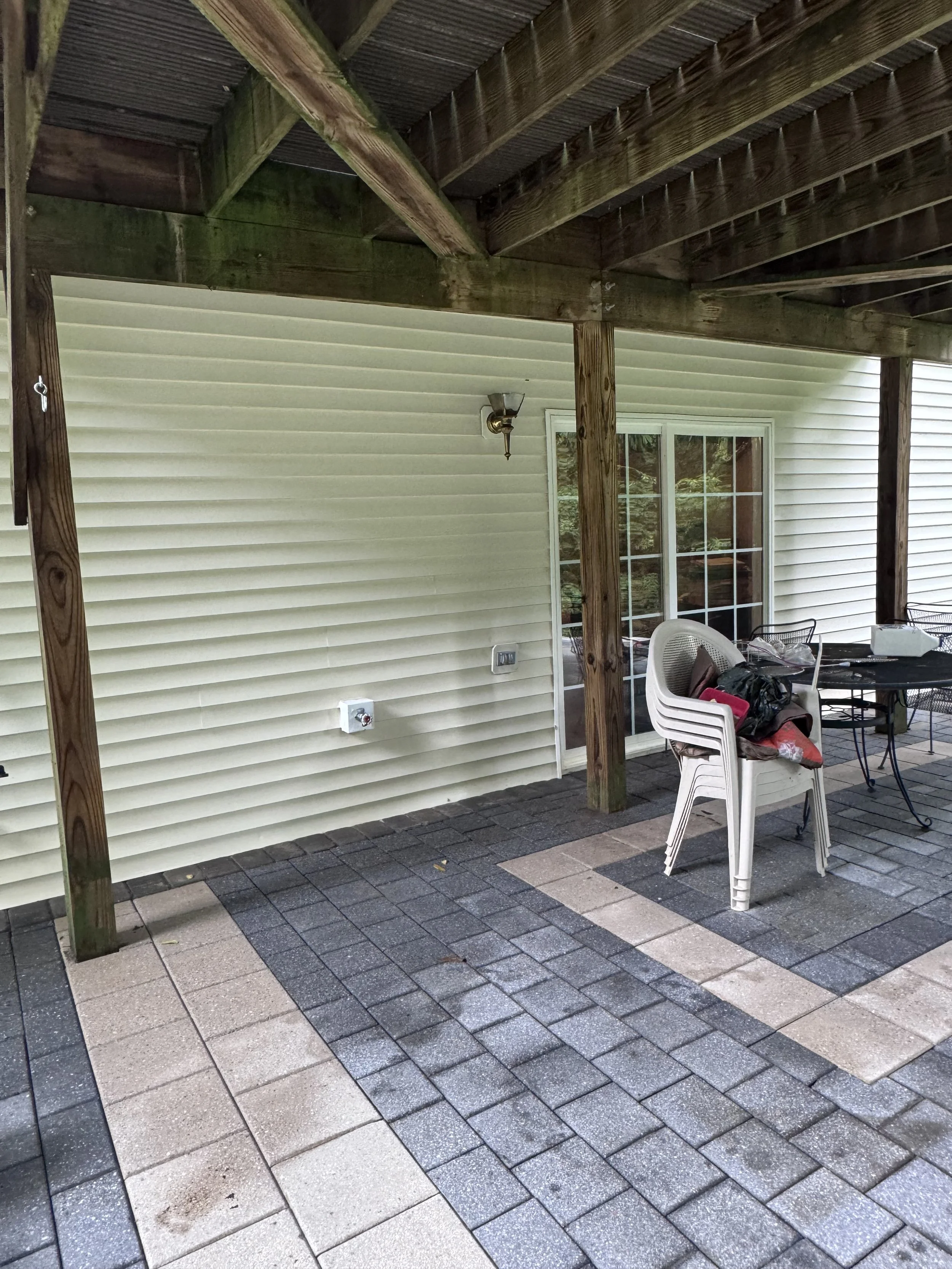 Back patio area with an overhead wooden patio cover, outdoor furniture including a stack of white plastic chairs, a black table, and a white chair with a pillow. The house's exterior has beige siding, a sliding glass door, and a wall-mounted outdoor 