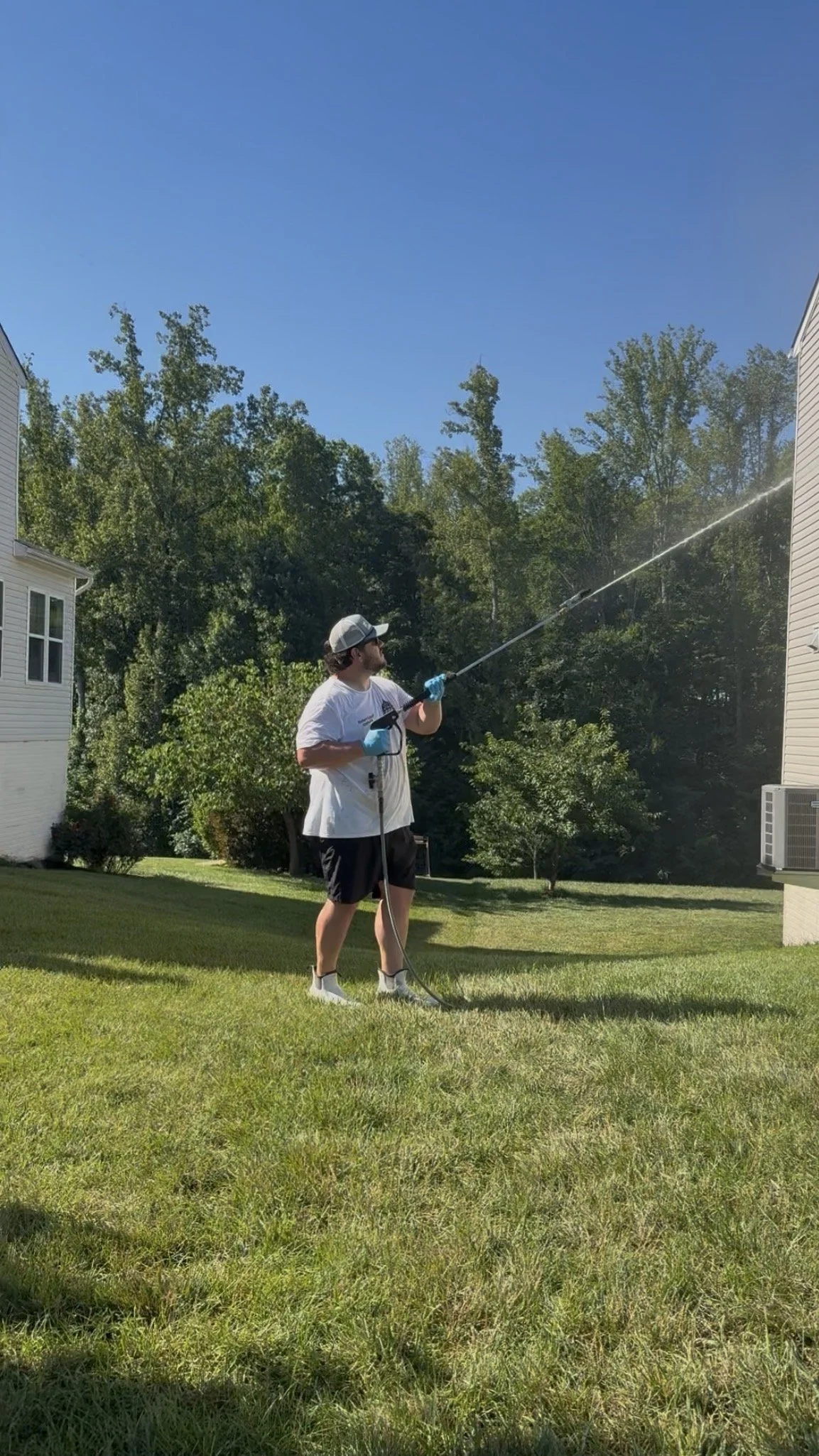 A man wearing a baseball cap, sunglasses, a white T-shirt, black shorts, and gloves is watering the lawn with a long hose in a backyard yard.