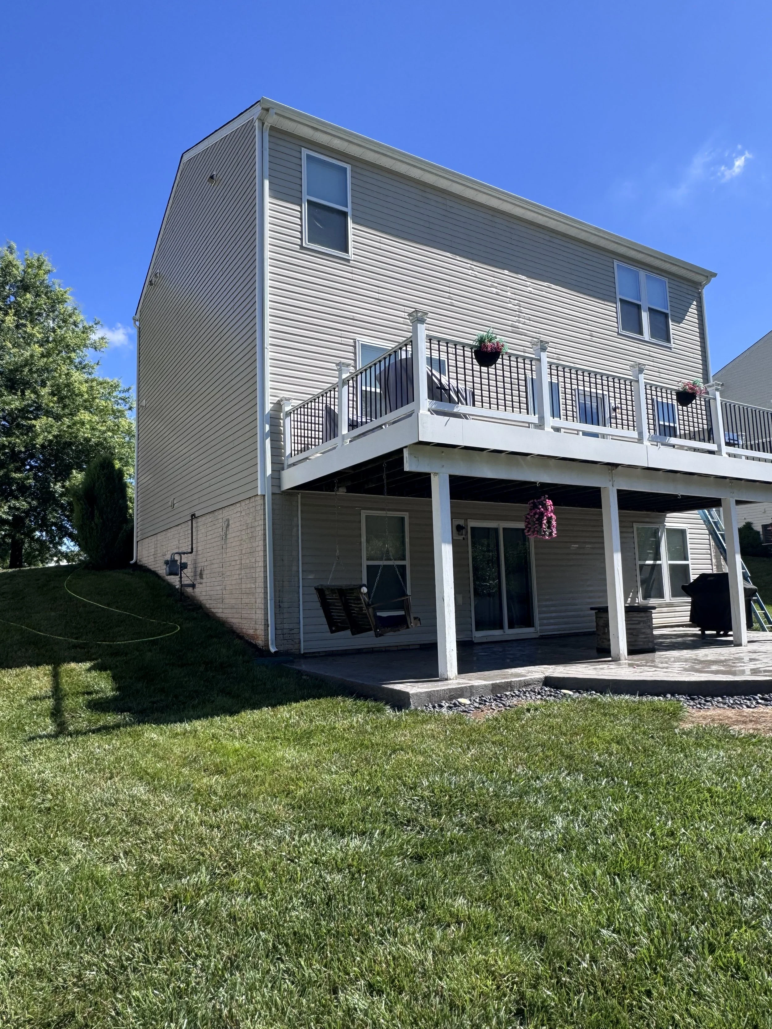 A multi-story house with beige siding, a deck with black railings and hanging flower baskets, large windows, and a grassy backyard under a blue sky.