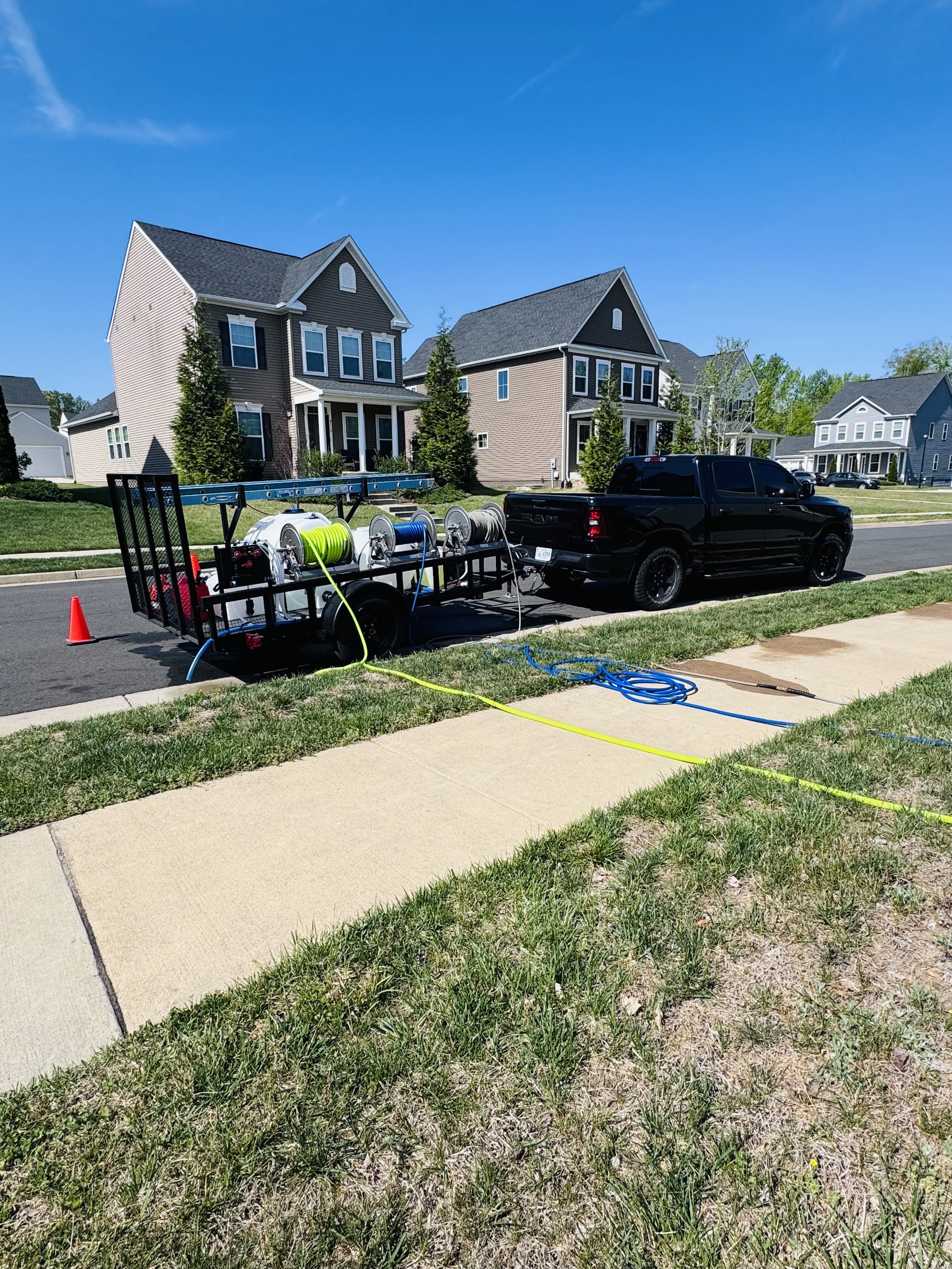 Exterior view of a neighborhood with two-story suburban houses, a black pickup truck parked on the street, and a trailer with water hoses and equipment, under a clear blue sky.