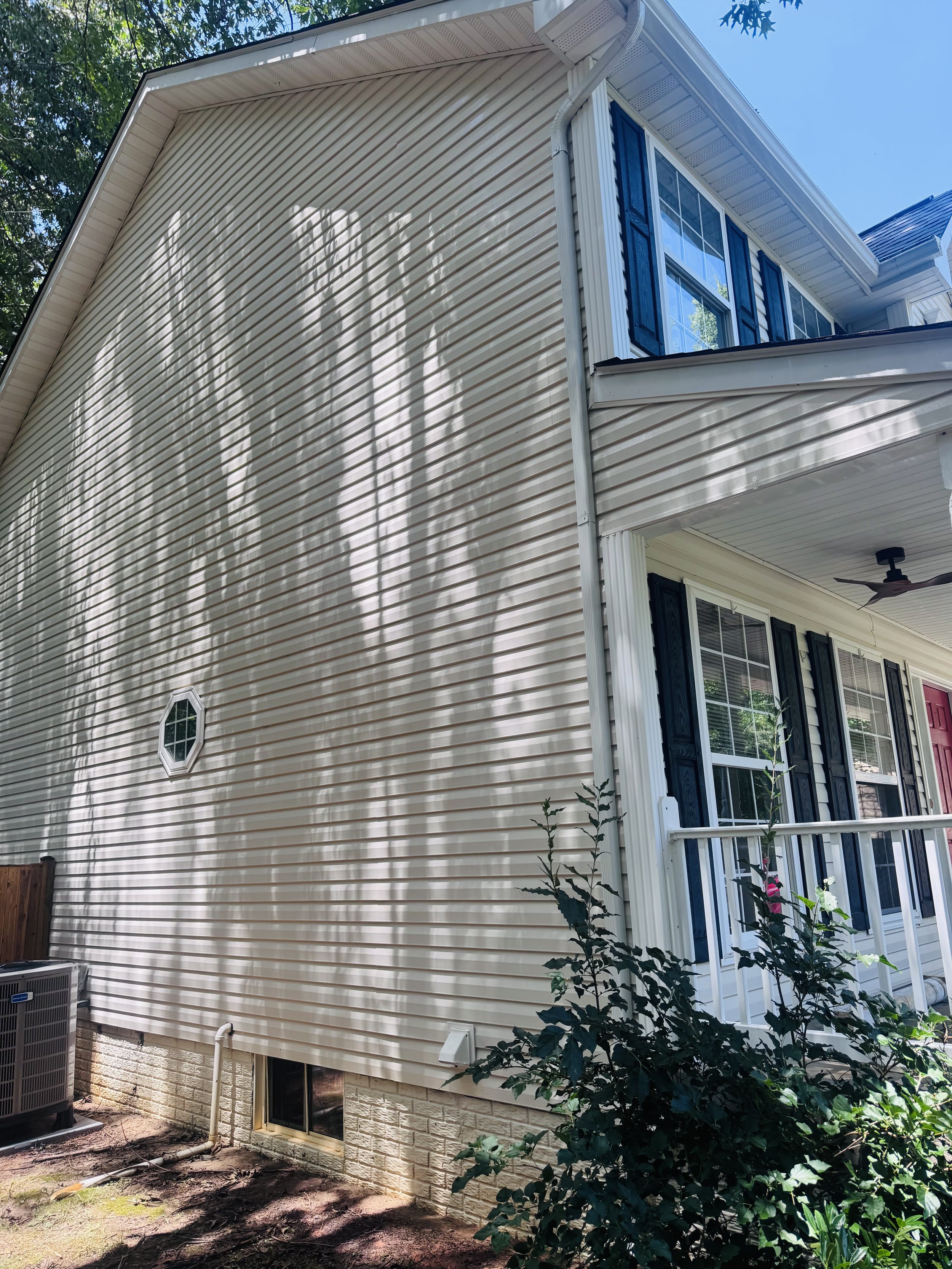 A beige house with blue shutters on windows, a white porch railing, and a ceiling fan. The house has metal siding and a small window near the foundation, with plants and bushes growing in front. The sky is clear and blue, with some tree shadows on th