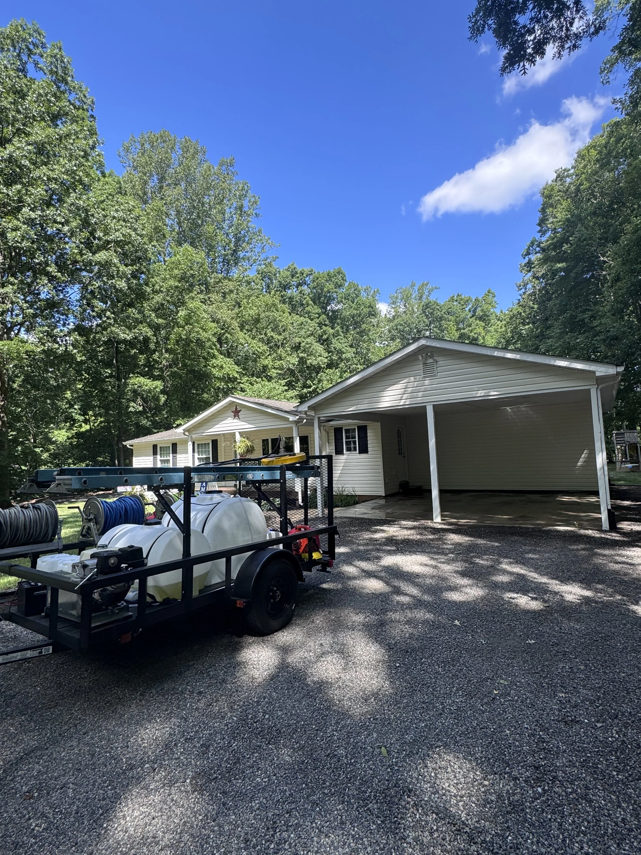 A suburban house with a porch and a red star decoration above the entrance, surrounded by tall green trees under a bright blue sky with some white clouds. In front of the house, there is a utility trailer loaded with hoses and tanks, parked on a grav