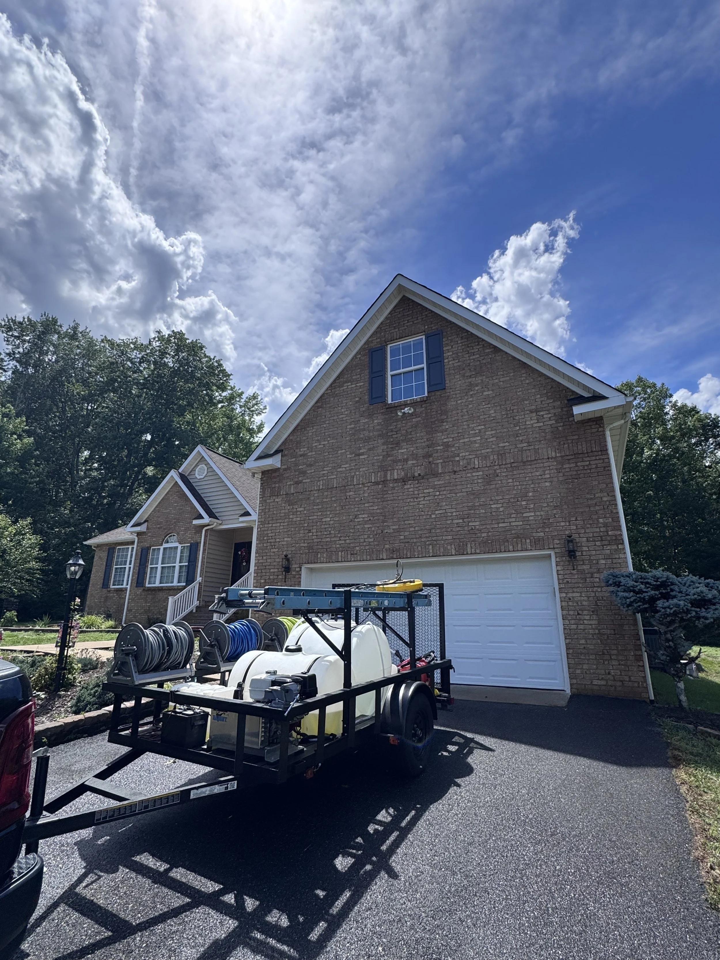 A suburban house with a brick exterior and a gabled roof, featuring a second-story window with shutters. A landscaping trailer with hoses and equipment is parked in the driveway. The sky is partly cloudy with patches of blue, and leafy trees surround
