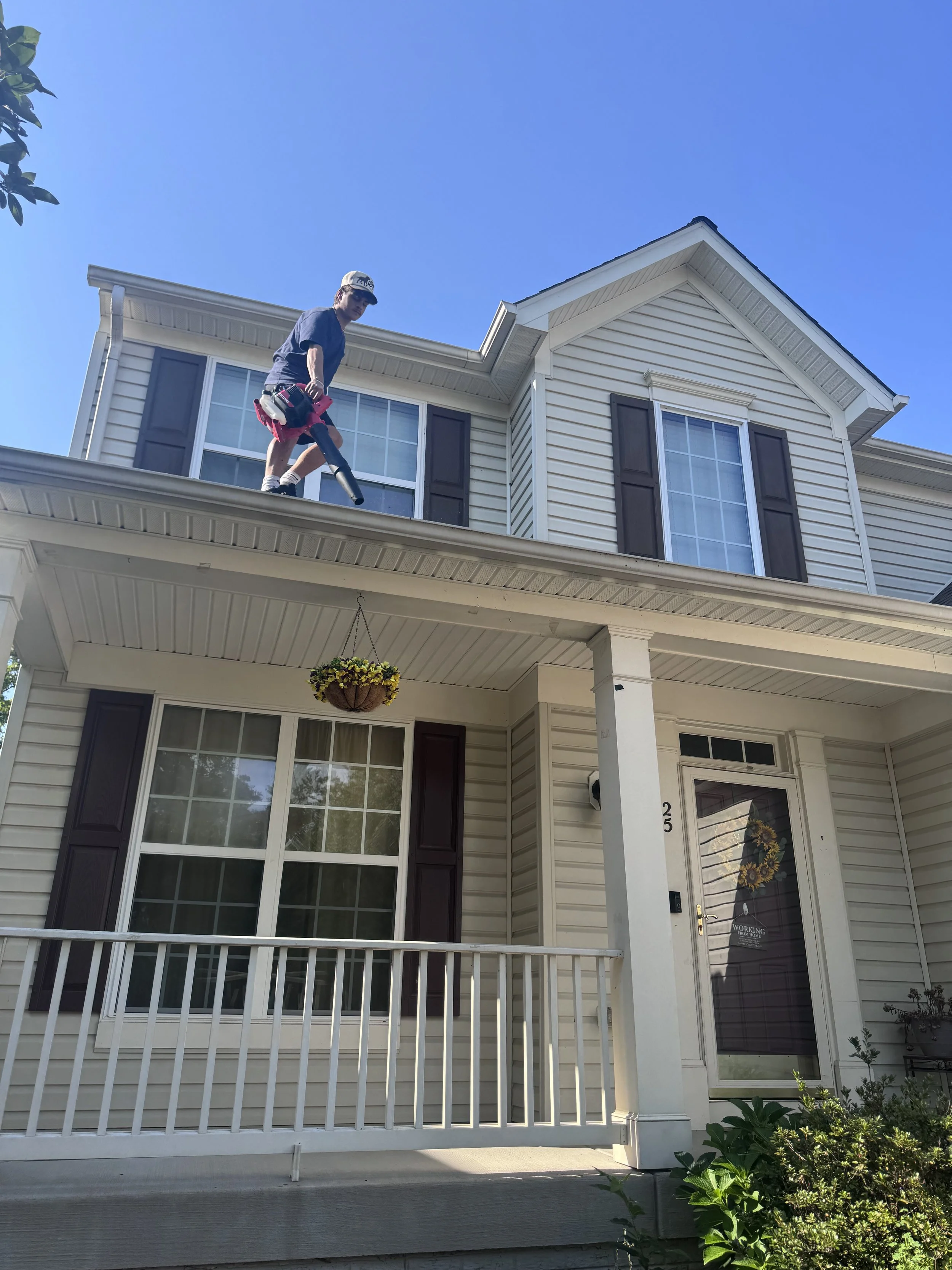 A man in a cap and casual clothes is standing on the roof of a house, holding a leaf blower, under a clear blue sky. The house has white siding, dark shutters, and a front porch with a hanging flower basket.