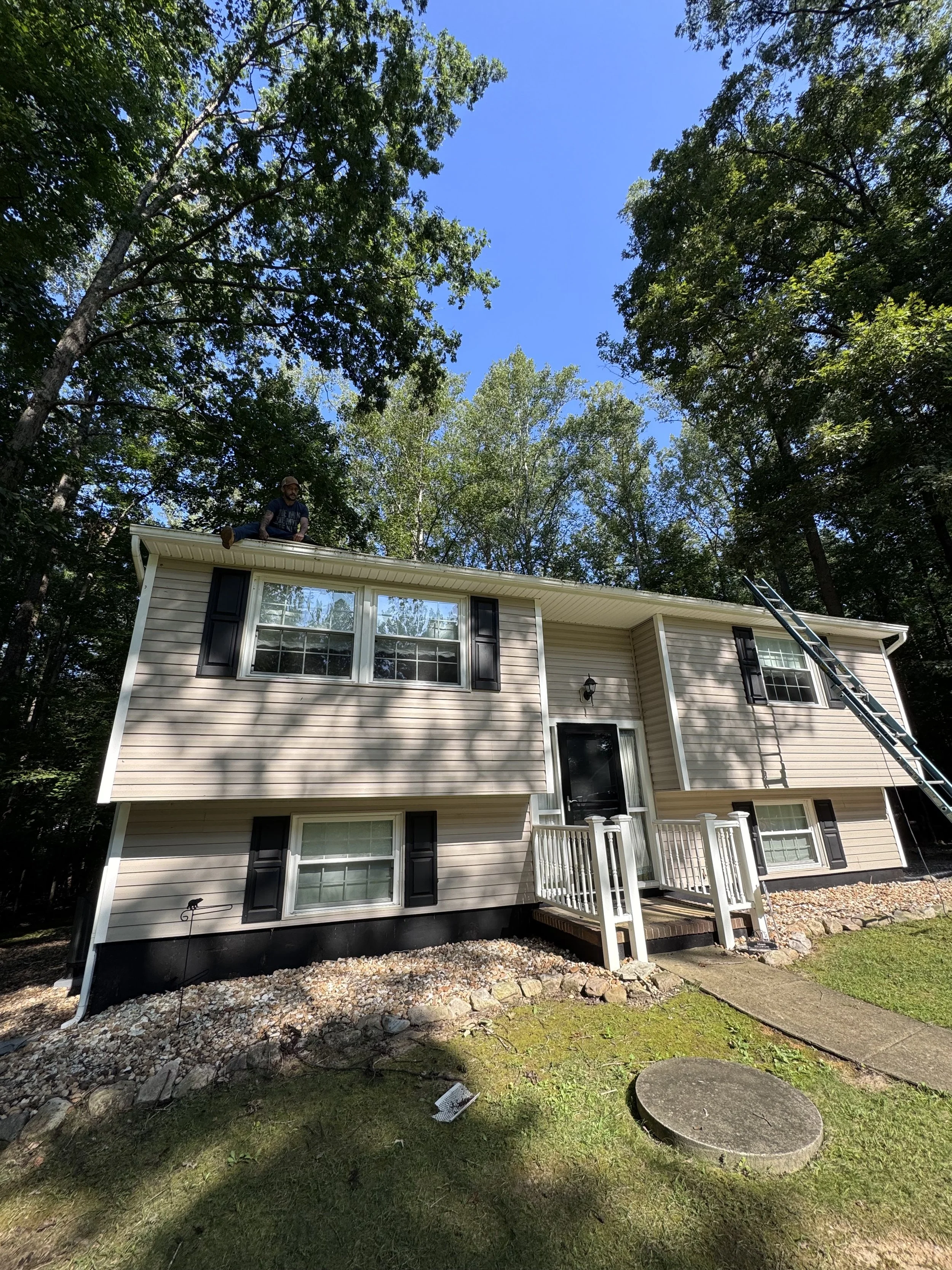 Two-story house with beige siding, black shutters, and a white porch railing. A man is crouching on the roof, working with tools. A ladder leans against the house, and there are trees with green leaves and a clear blue sky.