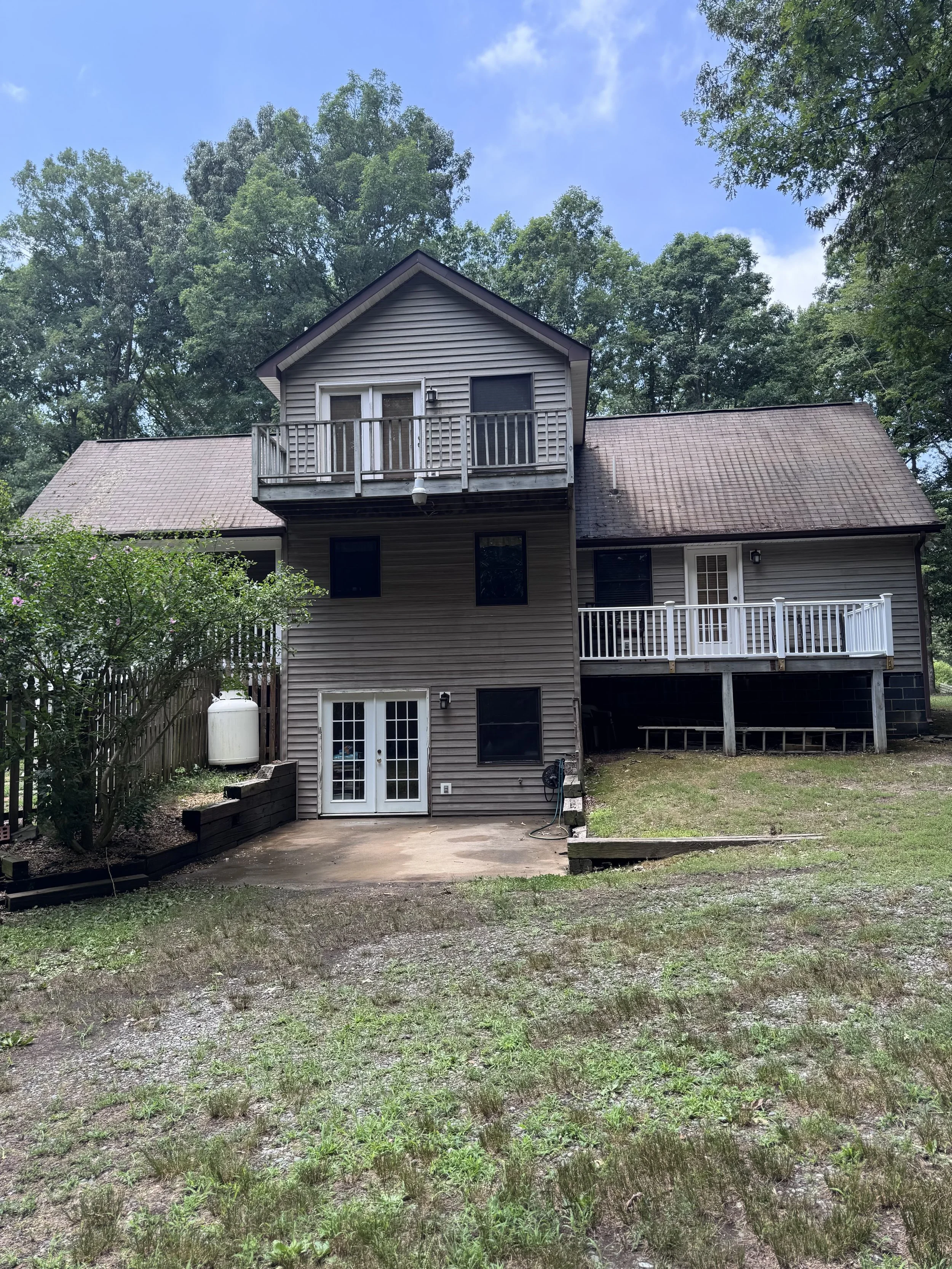 Backyard view of gray house with a small balcony, French doors, and a backyard patio area surrounded by trees and grass.