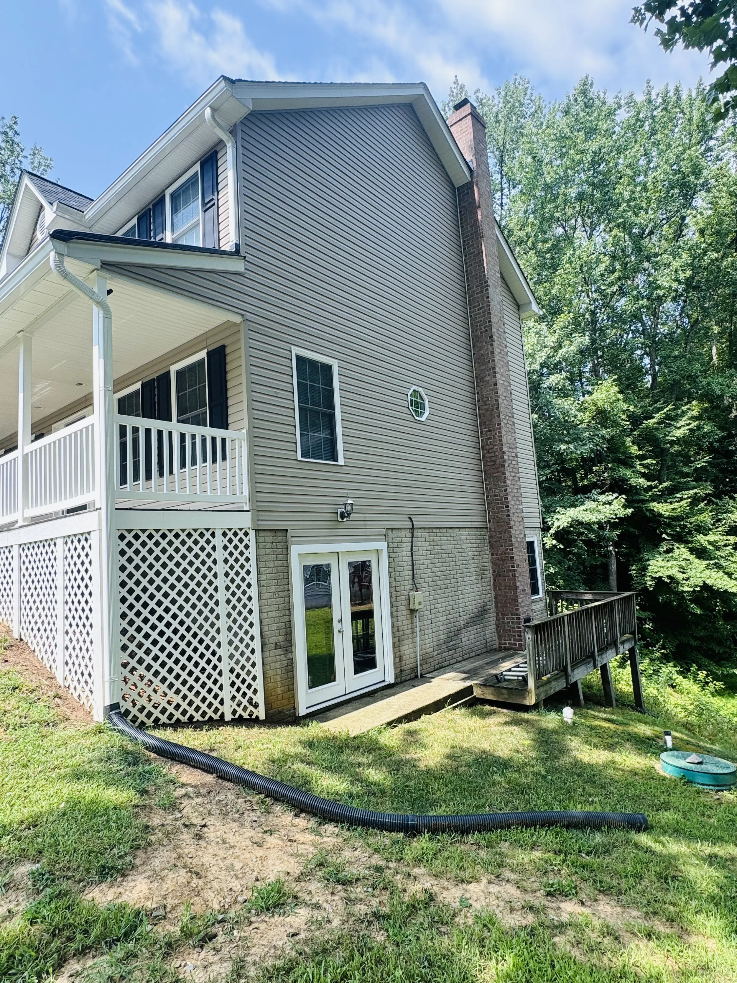 Backyard of a house with beige vinyl siding, a brick chimney, a white lattice privacy wall, and a wooden deck with stairs, surrounded by green grass and dense trees.