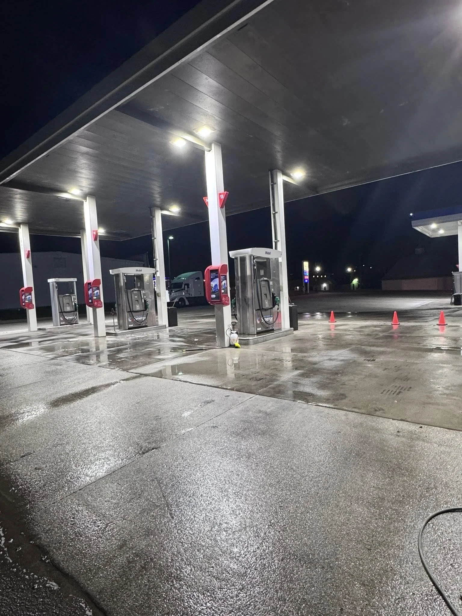 Nighttime scene at a gas station with illuminated fuel dispensers, wet pavement, orange traffic cones, and a dark sky.