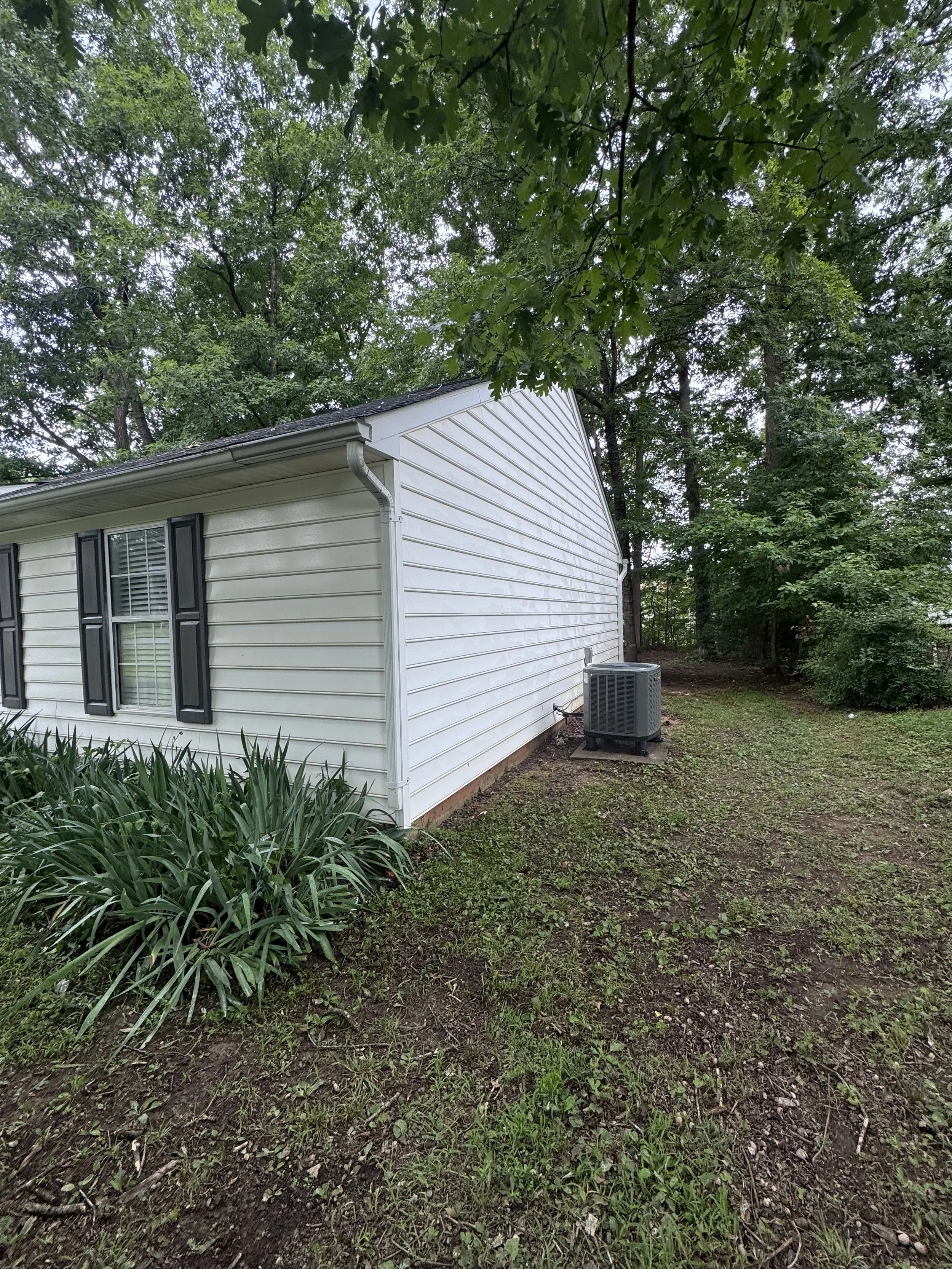 Side view of a white house with black shutters surrounded by green trees and grass, with an air conditioning unit outside.