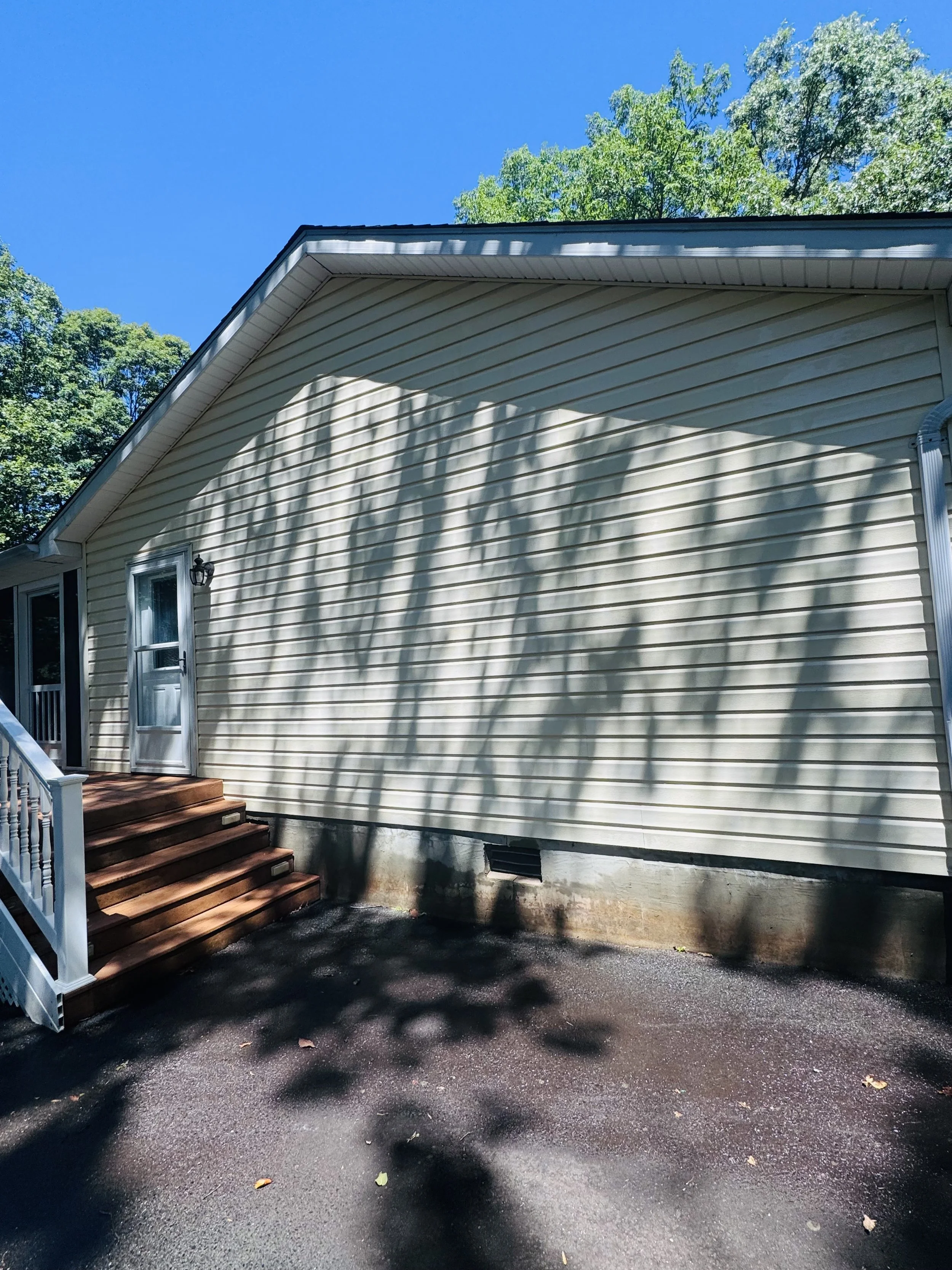 Backyard scene showing the side of a beige house with vinyl siding, a small window, and a black outdoor light fixture. There are wooden stairs leading to a door, and shadows of tree branches are cast on the house. The ground is wet, and the sky is bl