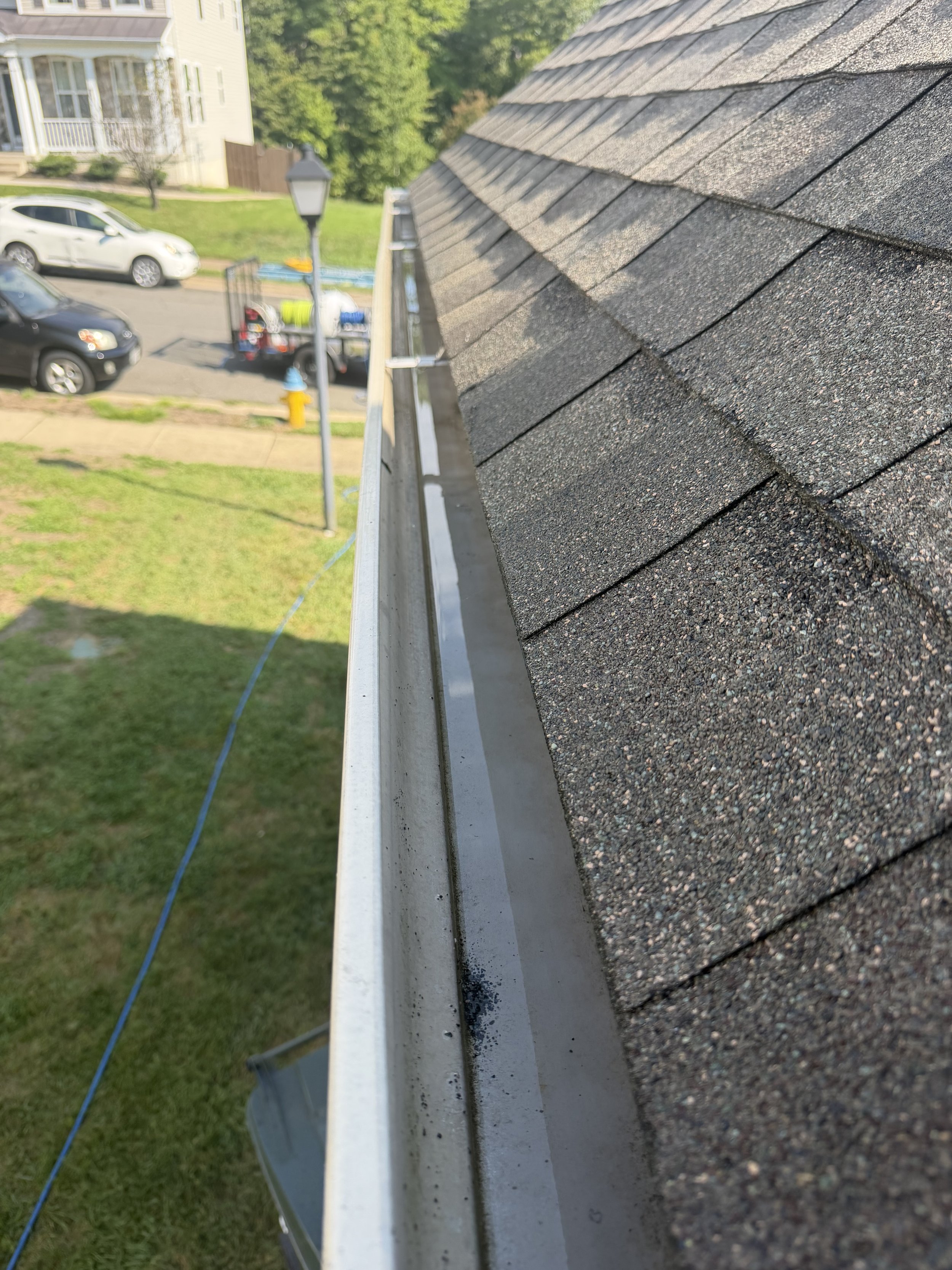 Close-up of a roof's edge with asphalt shingles and a gutter, overlooking a residential neighborhood with parked cars, a street lamp, a fire hydrant, and grass.