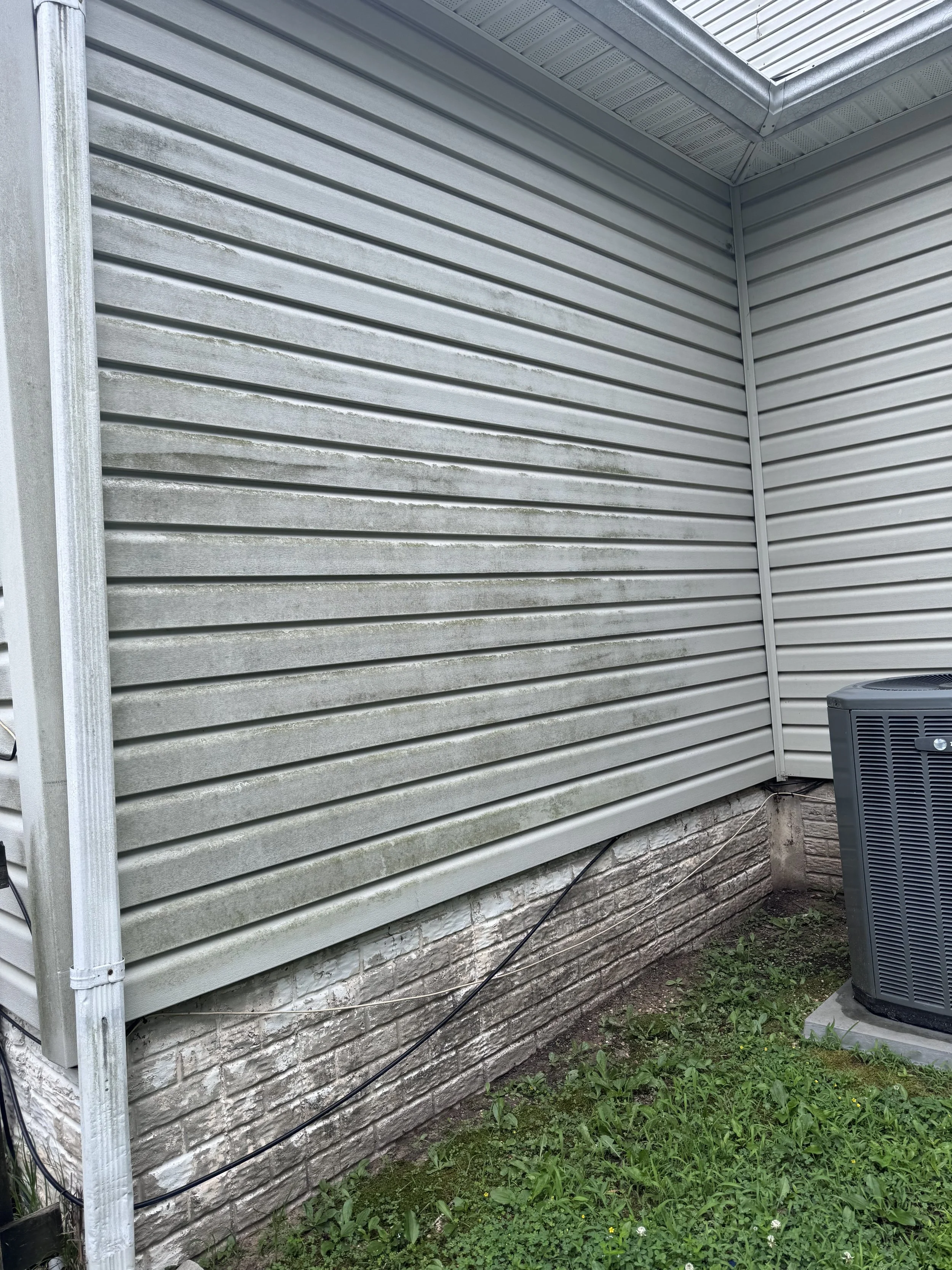 Side of a house with horizontal gray siding, a brick foundation, a small patch of grass and weeds, and an outdoor air conditioning unit.
