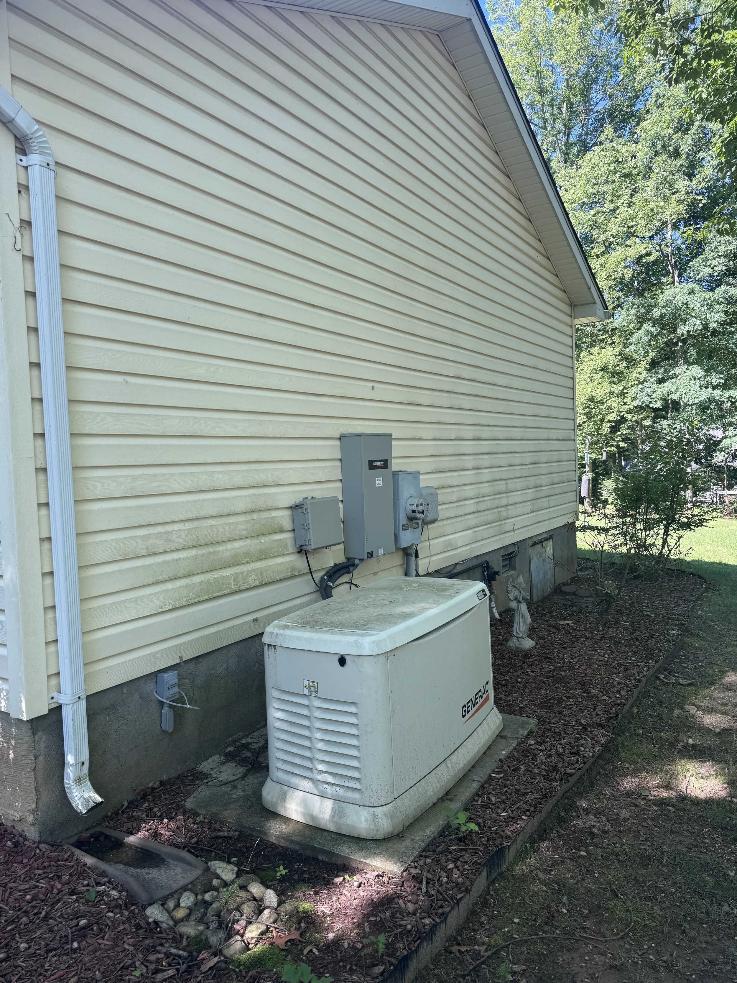 Backyard of a house with beige siding, outdoor electrical boxes, a generator, and a lawn with trees and bushes.