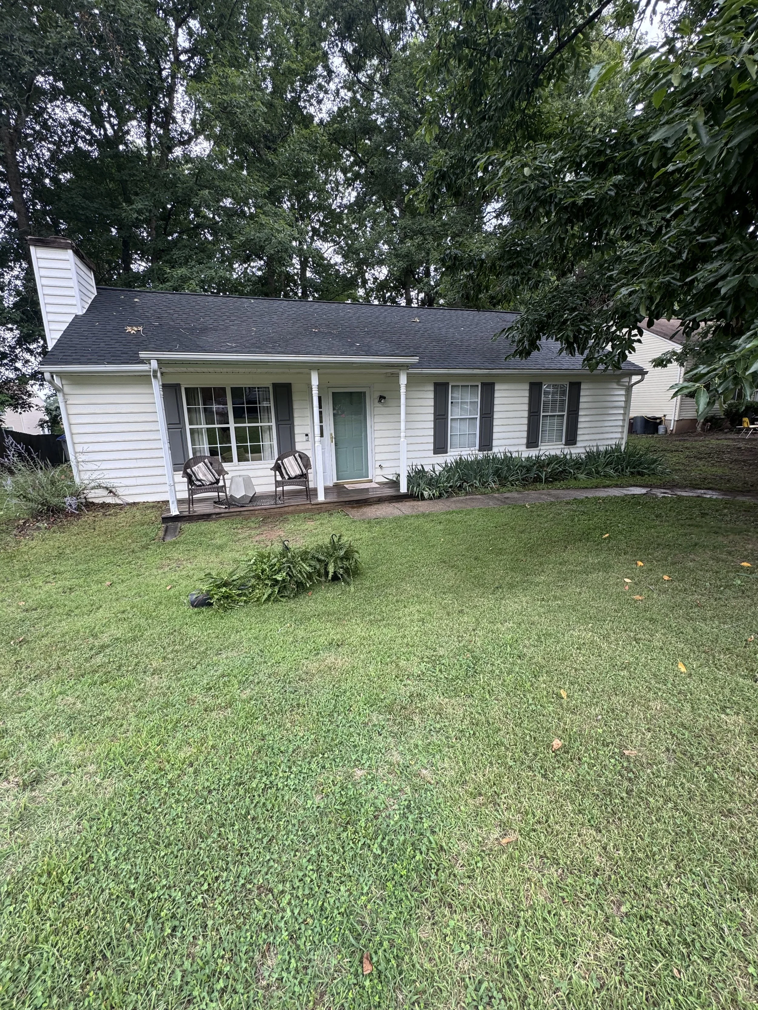 A white house with black shutters and a small front porch with two black chairs, set in a lush green yard with trees and bushes, during daylight.