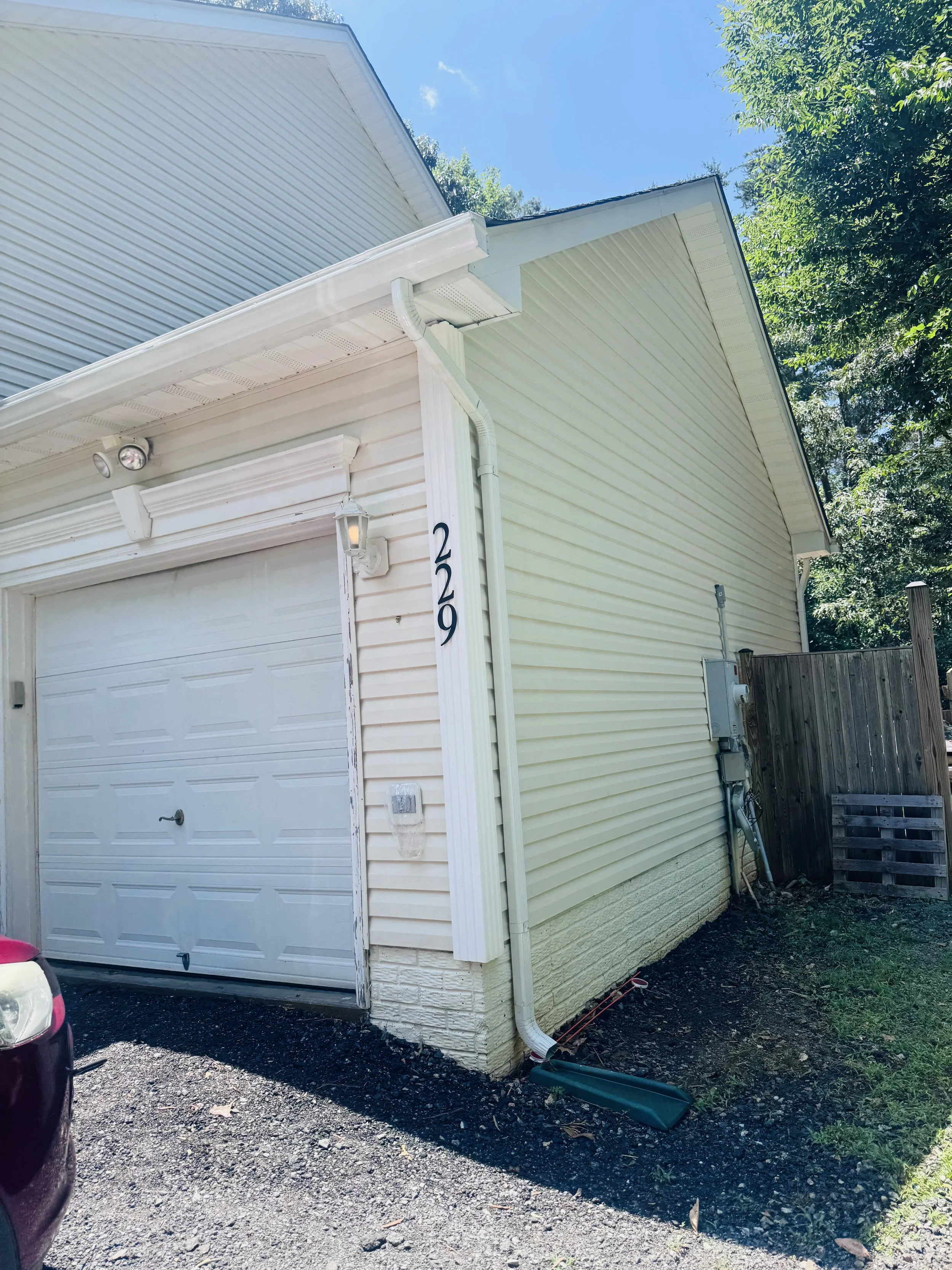 View of the side of a house with yellow siding, a white garage door, and a white gutter system. The house has a partial view of the street and parked vehicles. Trees and a clear blue sky are visible in the background.