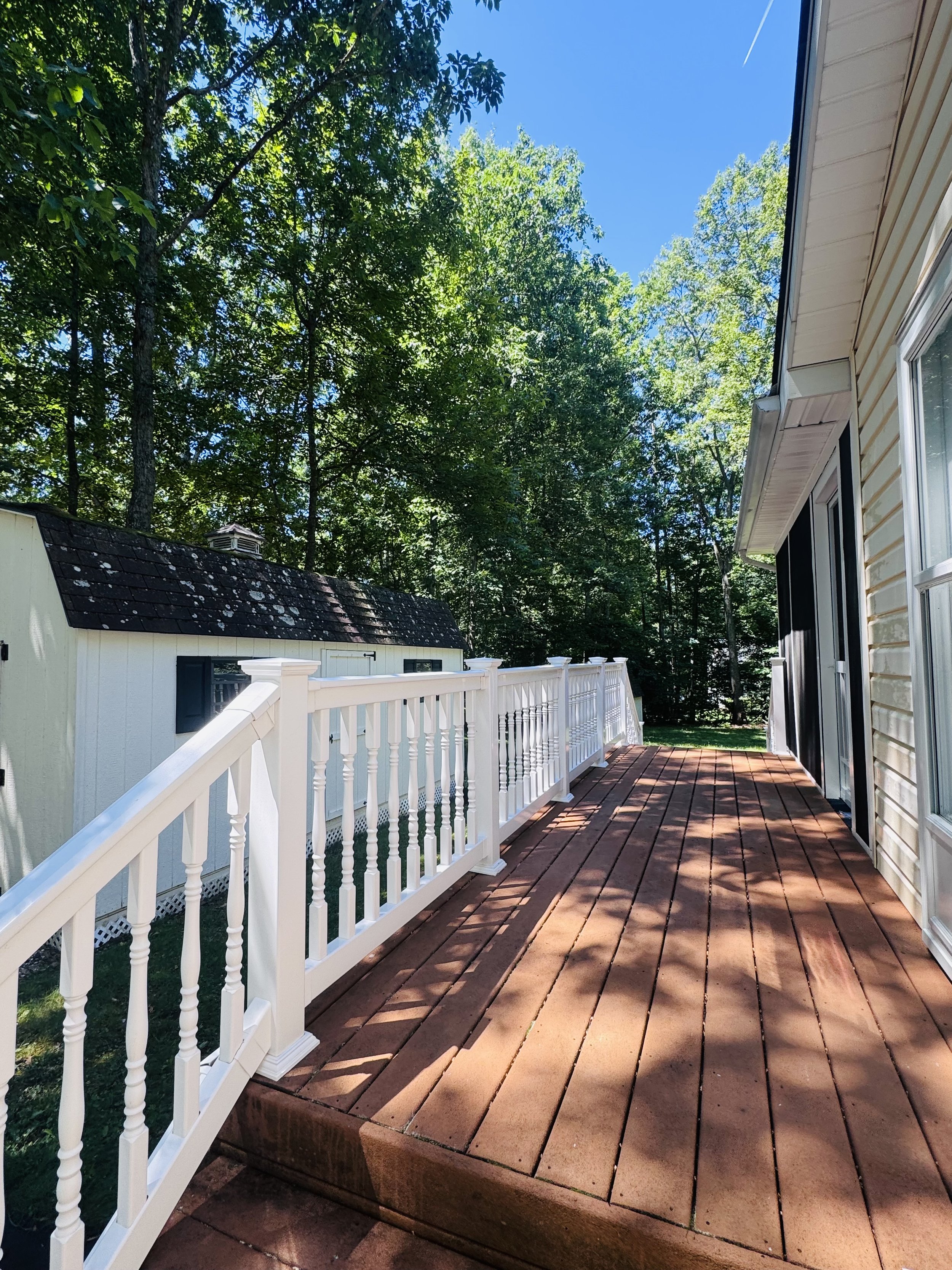 View of a backyard deck with reddish-brown wooden planks, white railing, and adjacent house siding, with trees and a bright blue sky in the background.