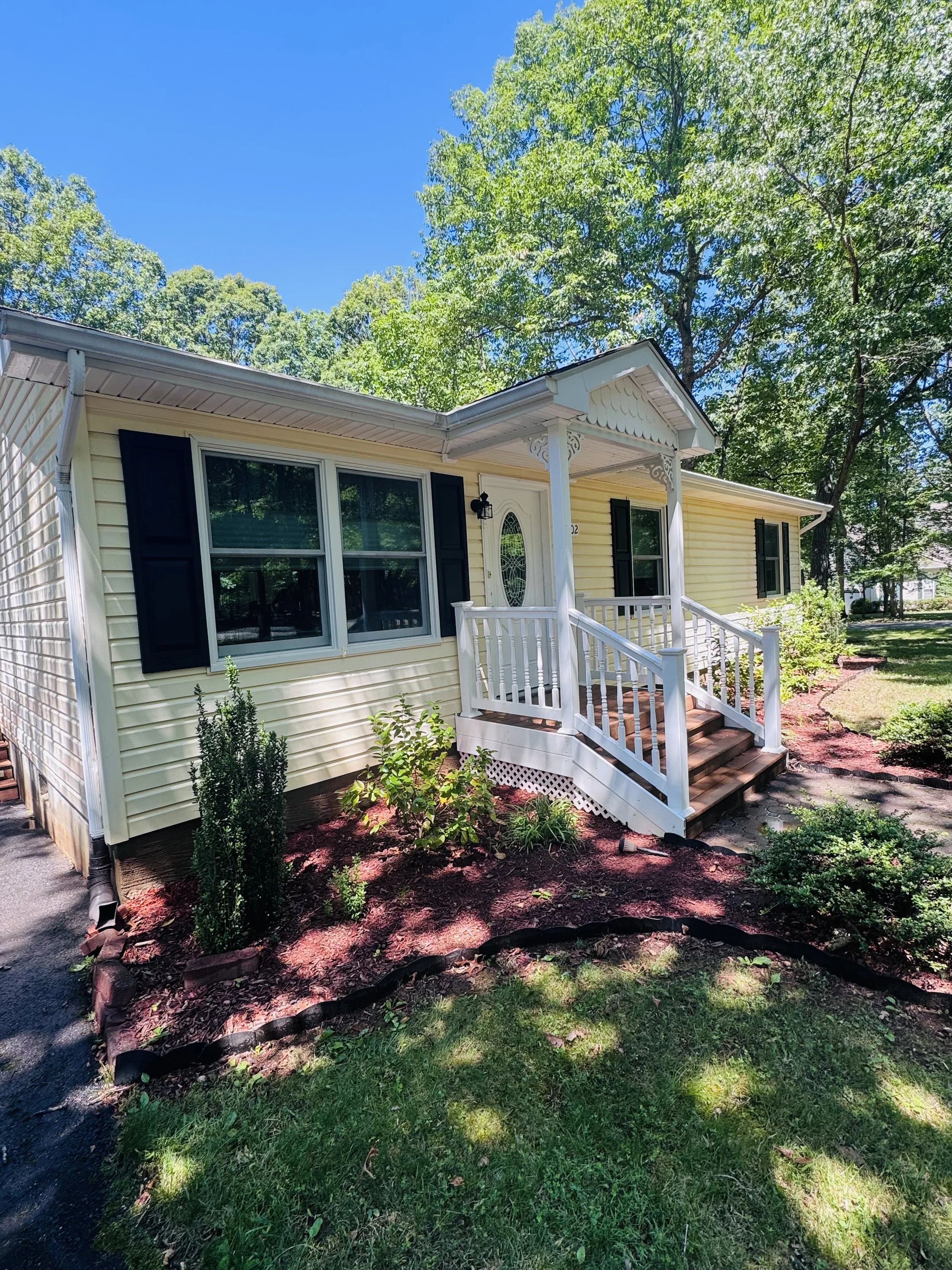 Front view of a yellow house with black shutters, white trim, a small front porch with white railing, surrounded by a garden with plants and mulch, under a clear blue sky and green trees.