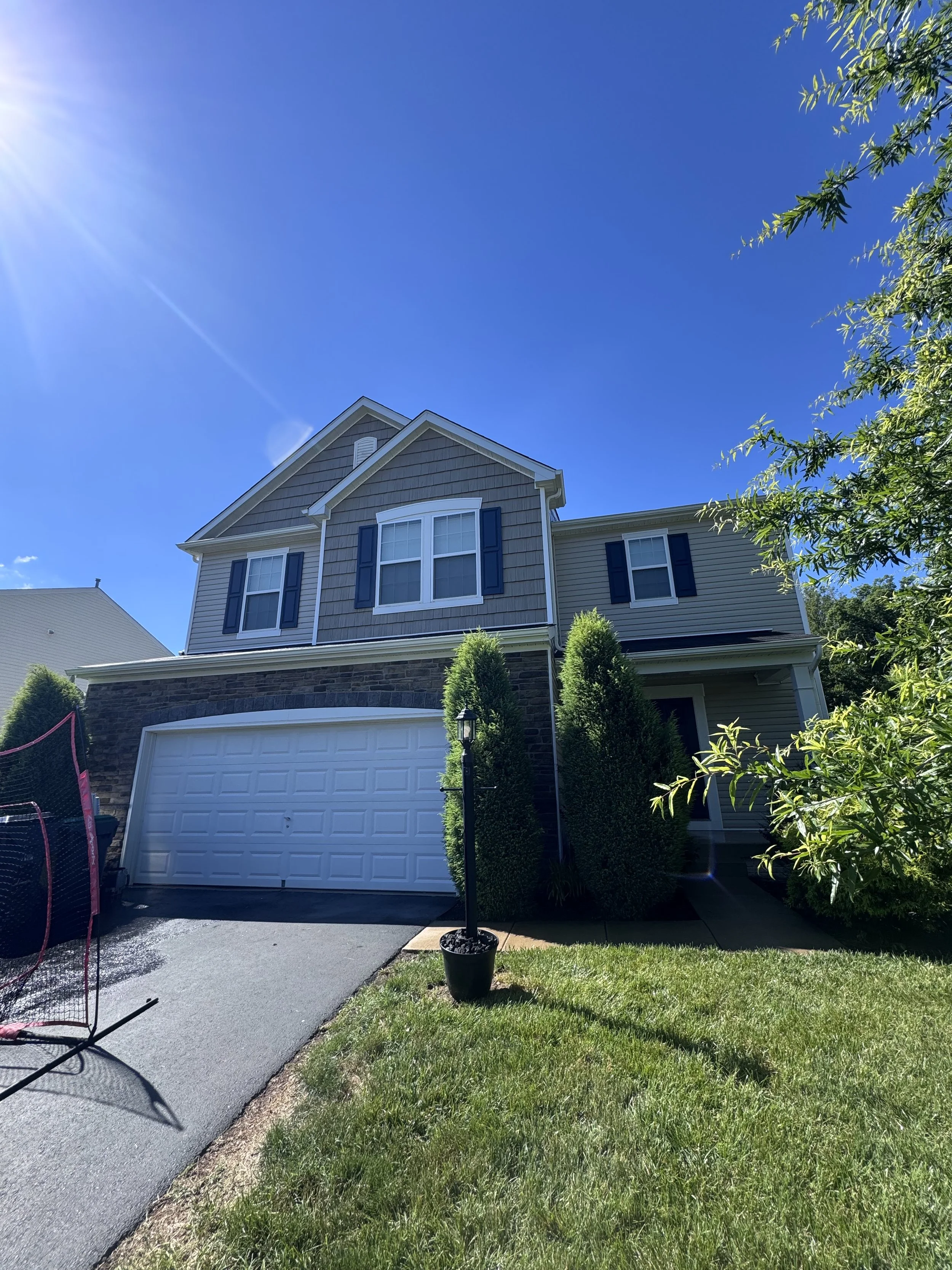 Front yard view of a two-story house with a white garage door, grey siding, and black shutters, under a bright blue sky with sunlight, greenery, and a small tree in a pot.