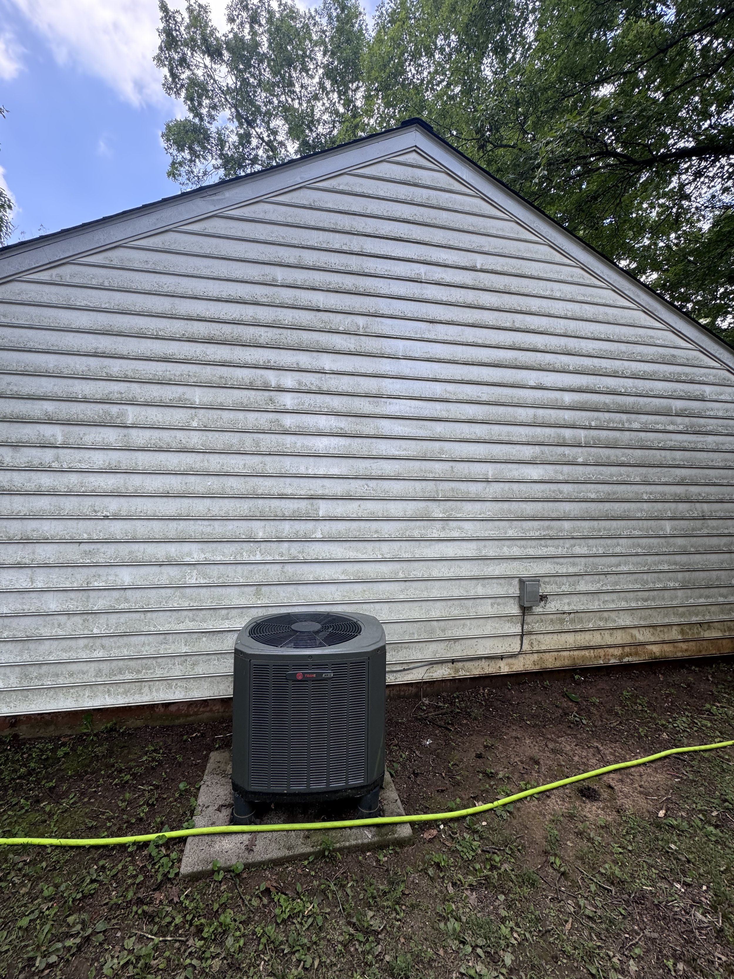 The image shows the side of a house with white siding that is dirty and stained. An outdoor air conditioning unit is placed on a concrete slab in the yard below, with a yellow garden hose and some patches of grass and dirt on the ground.
