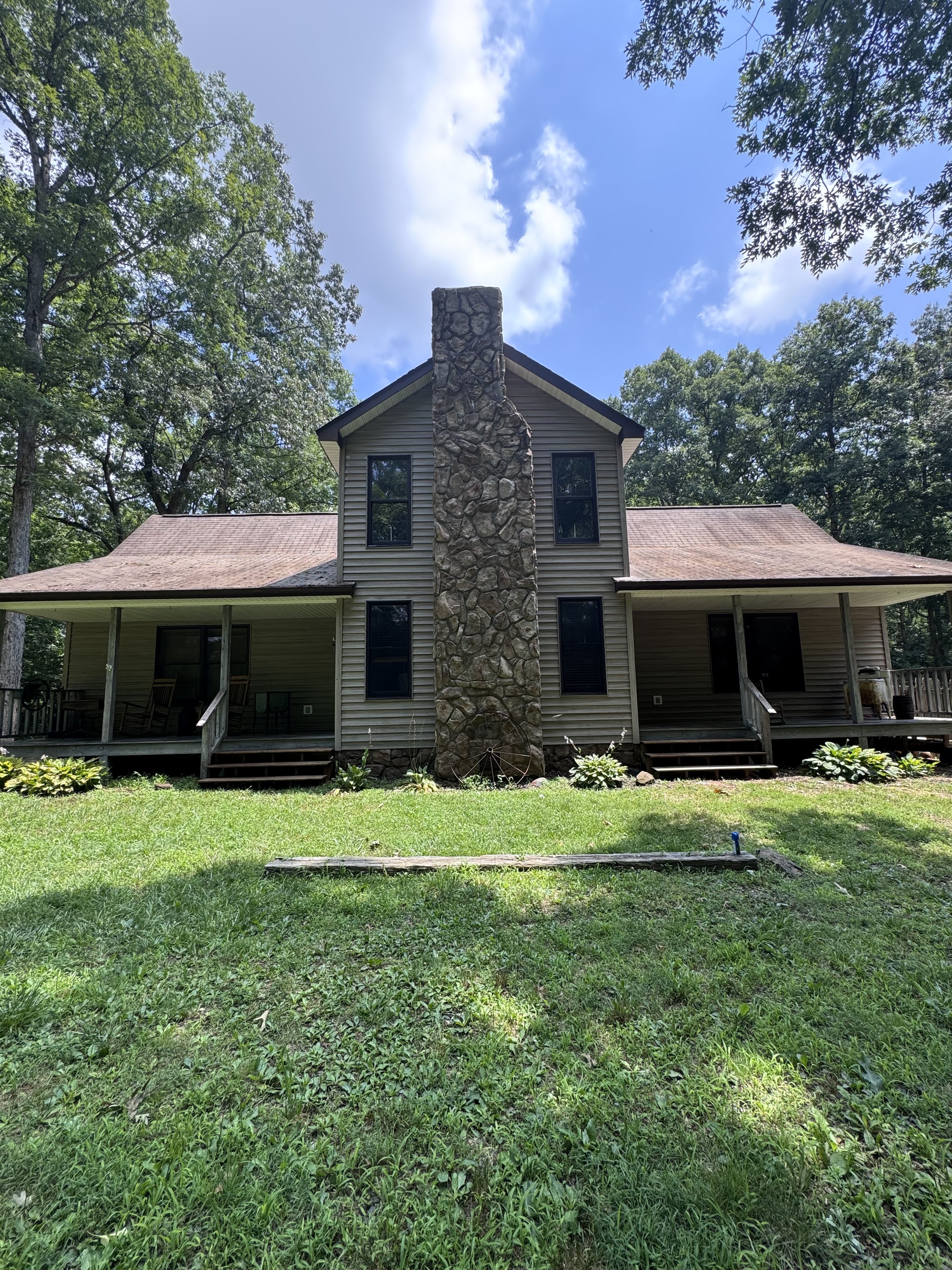 A house with beige siding and black framed windows, surrounded by trees and a grassy yard, with a large stone chimney at the center.