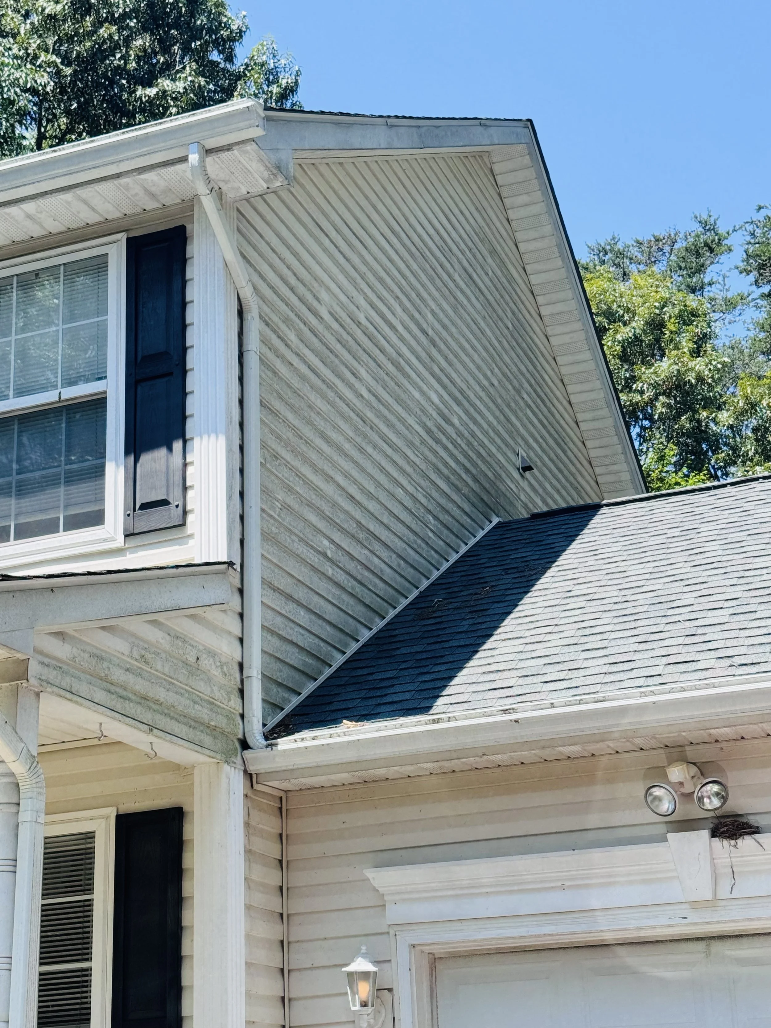 Close-up of the upper corner of a house with a white exterior, black shutters, a window with blinds, a gutter system, a rock wall, a patio light, and some trees in the background under a clear blue sky.