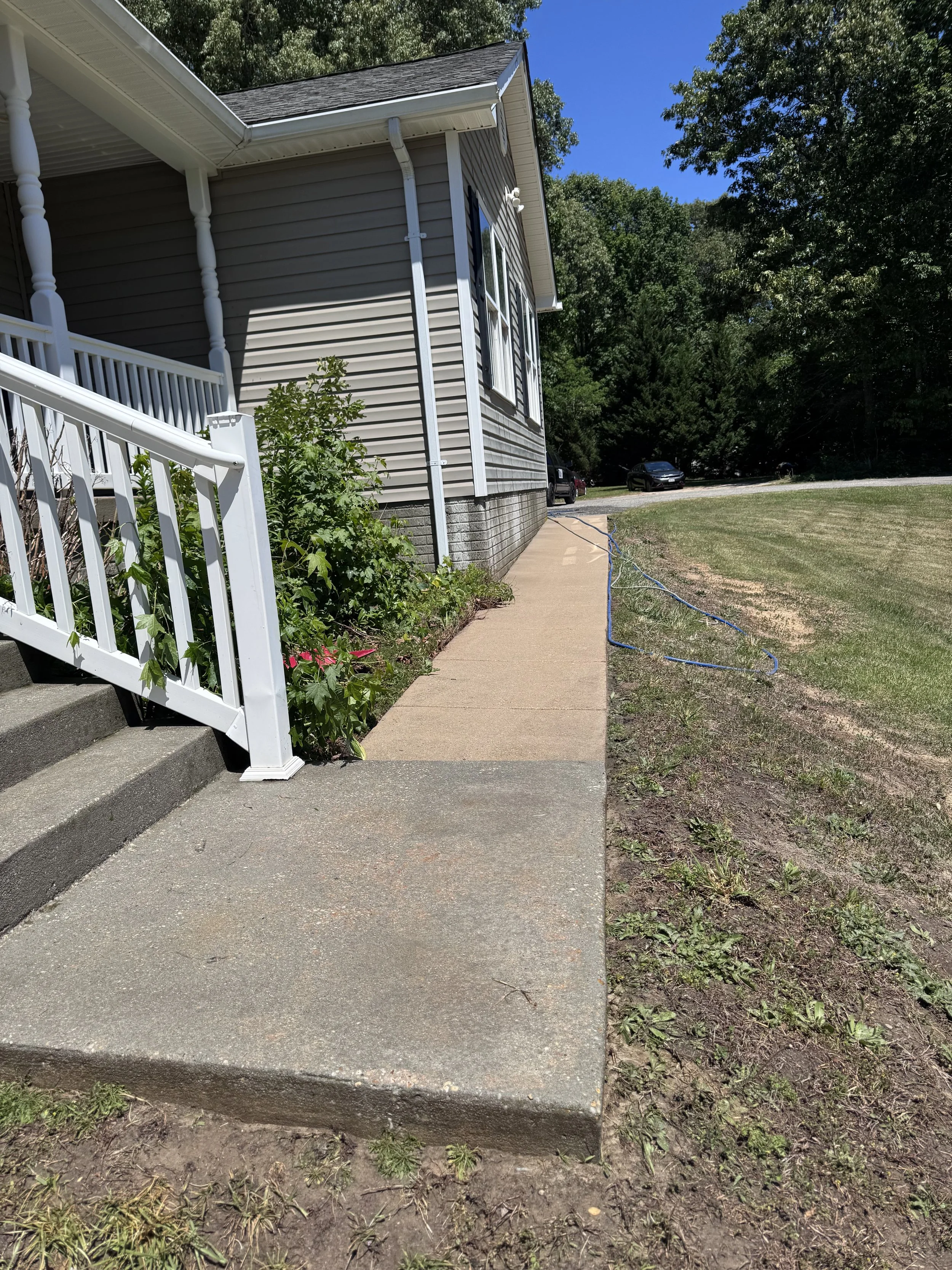 Side view of a house with beige siding, white trim, and a porch with white railing. A concrete walkway runs alongside the house with a garden bed and some plants. There are cars parked in the background on a gravel driveway. The yard has grass and so