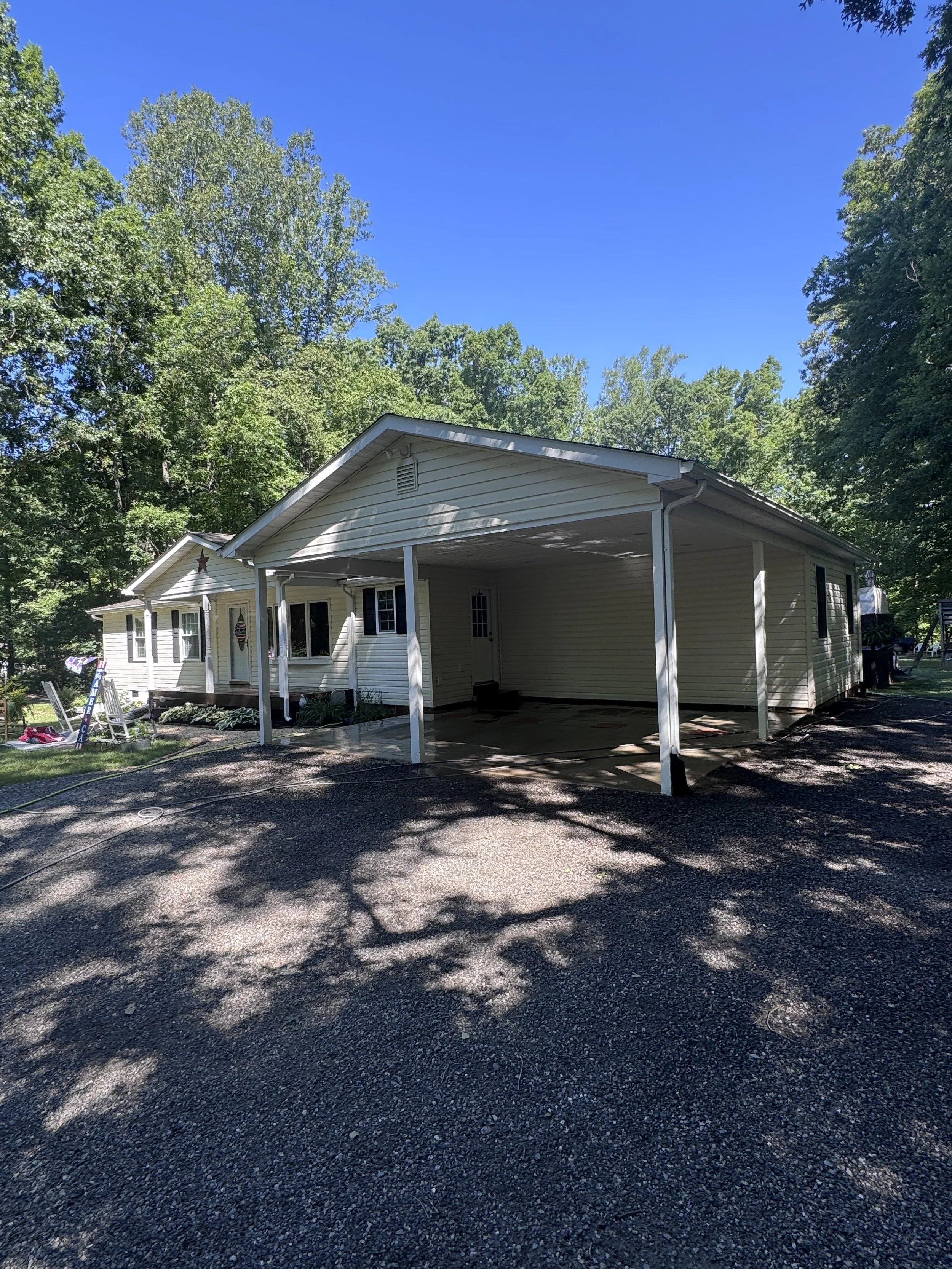 A house with white siding features a covered front porch with white pillars. There are two windows on the front and a door under the porch. The yard has a small garden area with plants and decorations, and the driveway is paved with asphalt. Tall gre