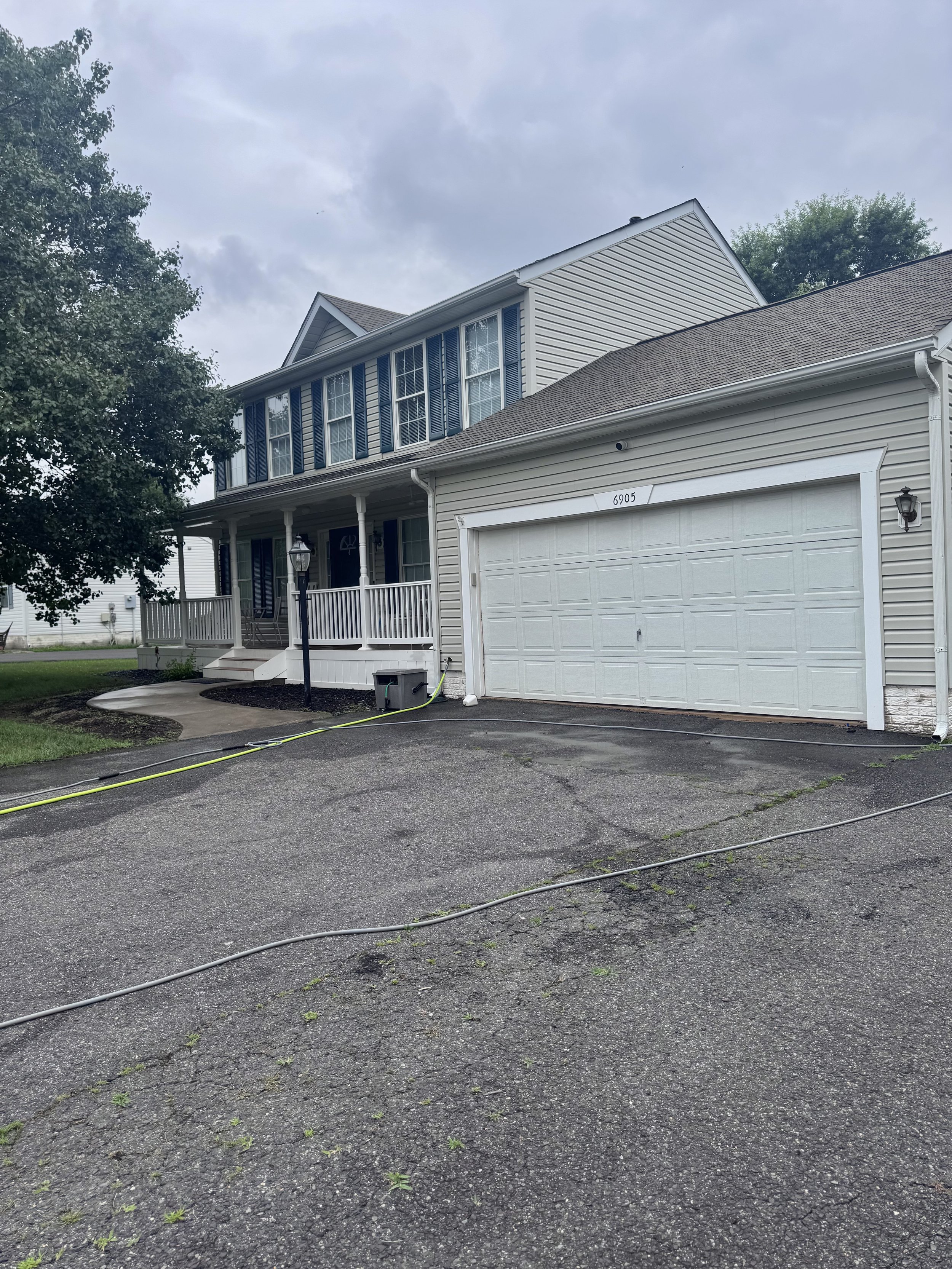 Front of a residential house with beige siding, white garage door, black shutters, a porch with white railing, and a tree to the left. The driveway has a hose and some small plants growing through the cracks.