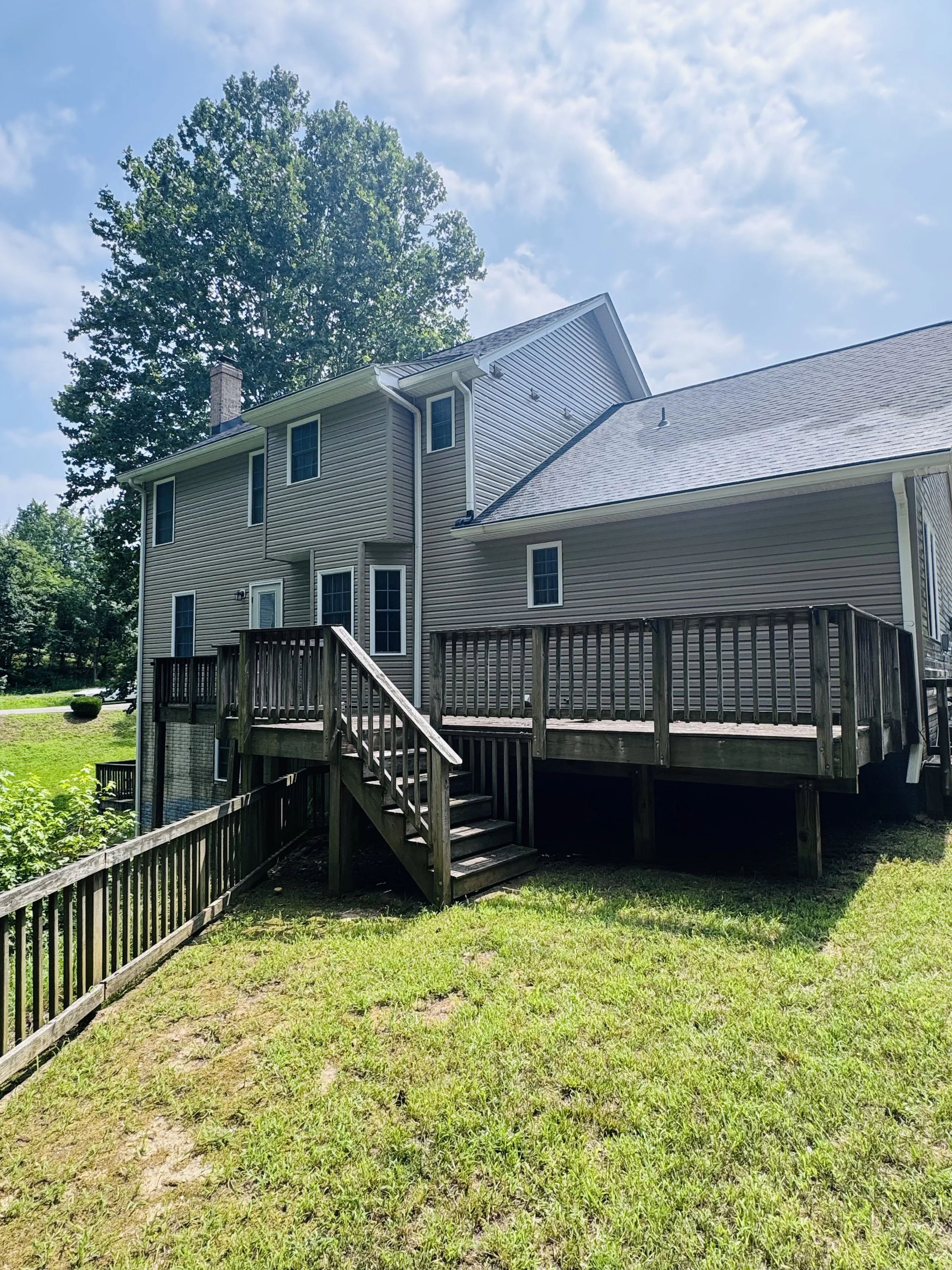 Backyard view of a multi-story house with beige siding, multiple windows, and a wooden deck with stairs, surrounded by green grass and trees under a partly cloudy sky.