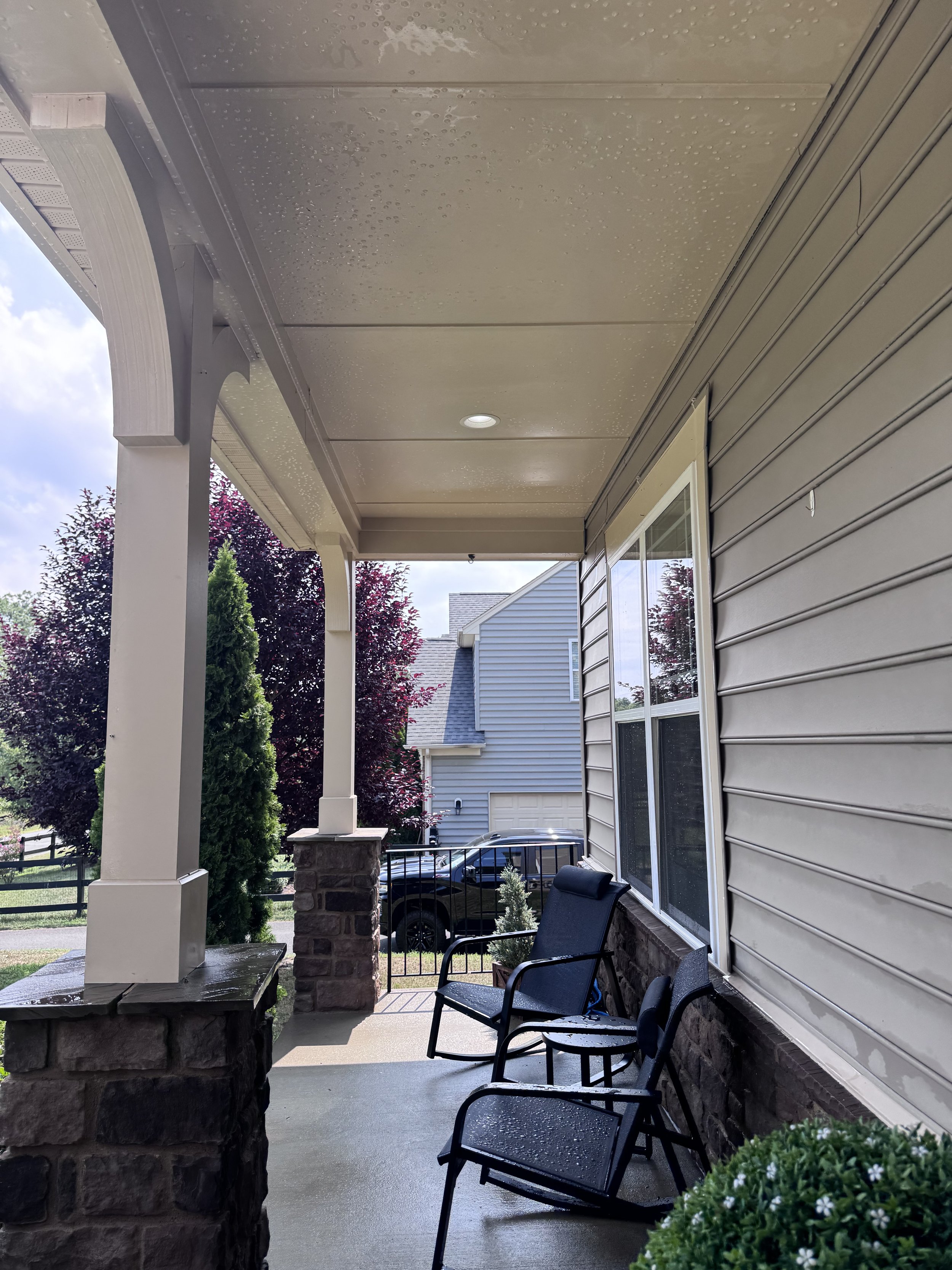 Covered front porch with a concrete floor, black chairs, a brick pillar, and neighboring houses and trees in the background, with raindrops on the ceiling.