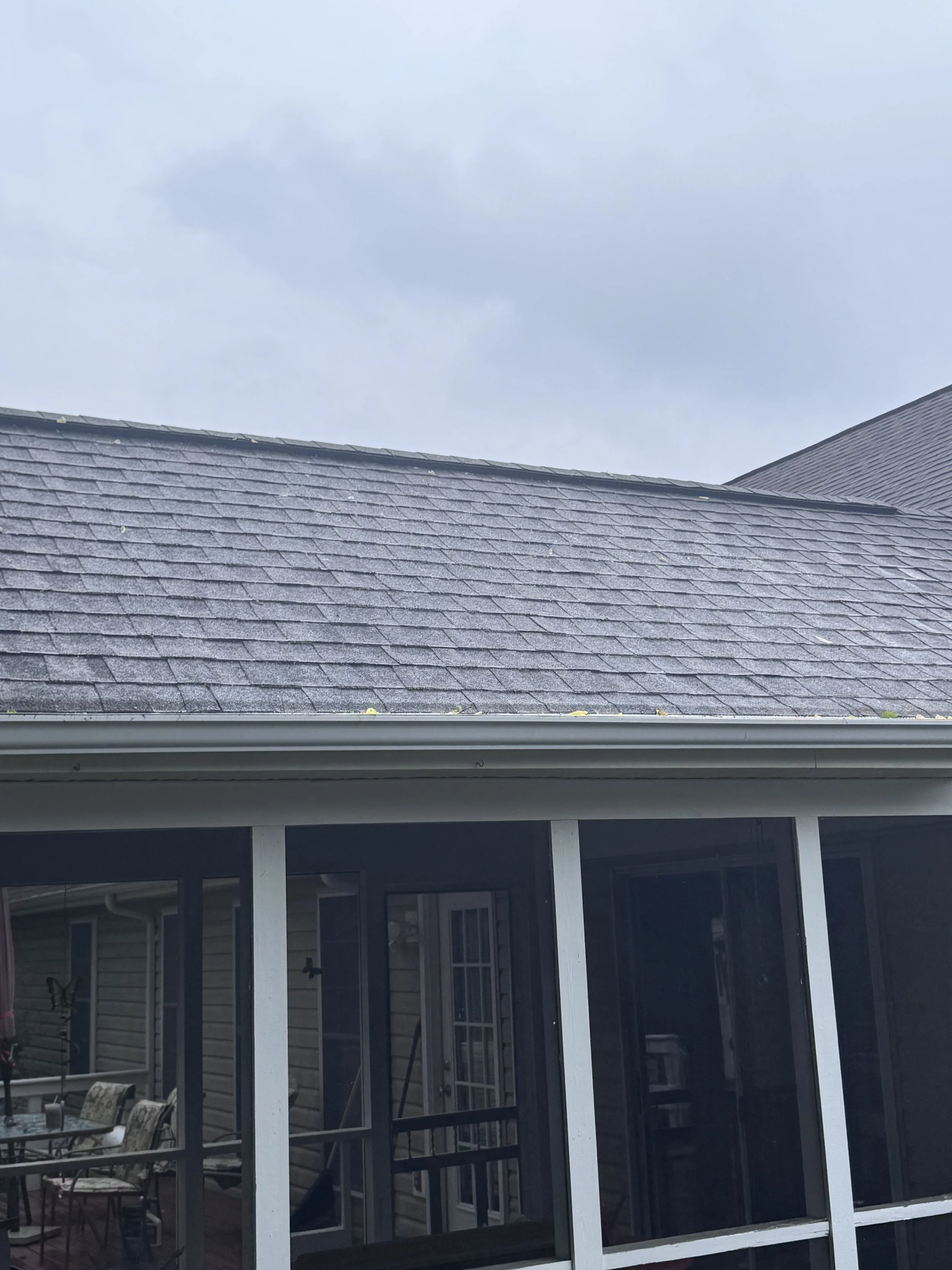 View of a house roof made of gray shingles, viewed from a windowed porch area with white framing, with an outdoor patio and cloudy sky in the background.