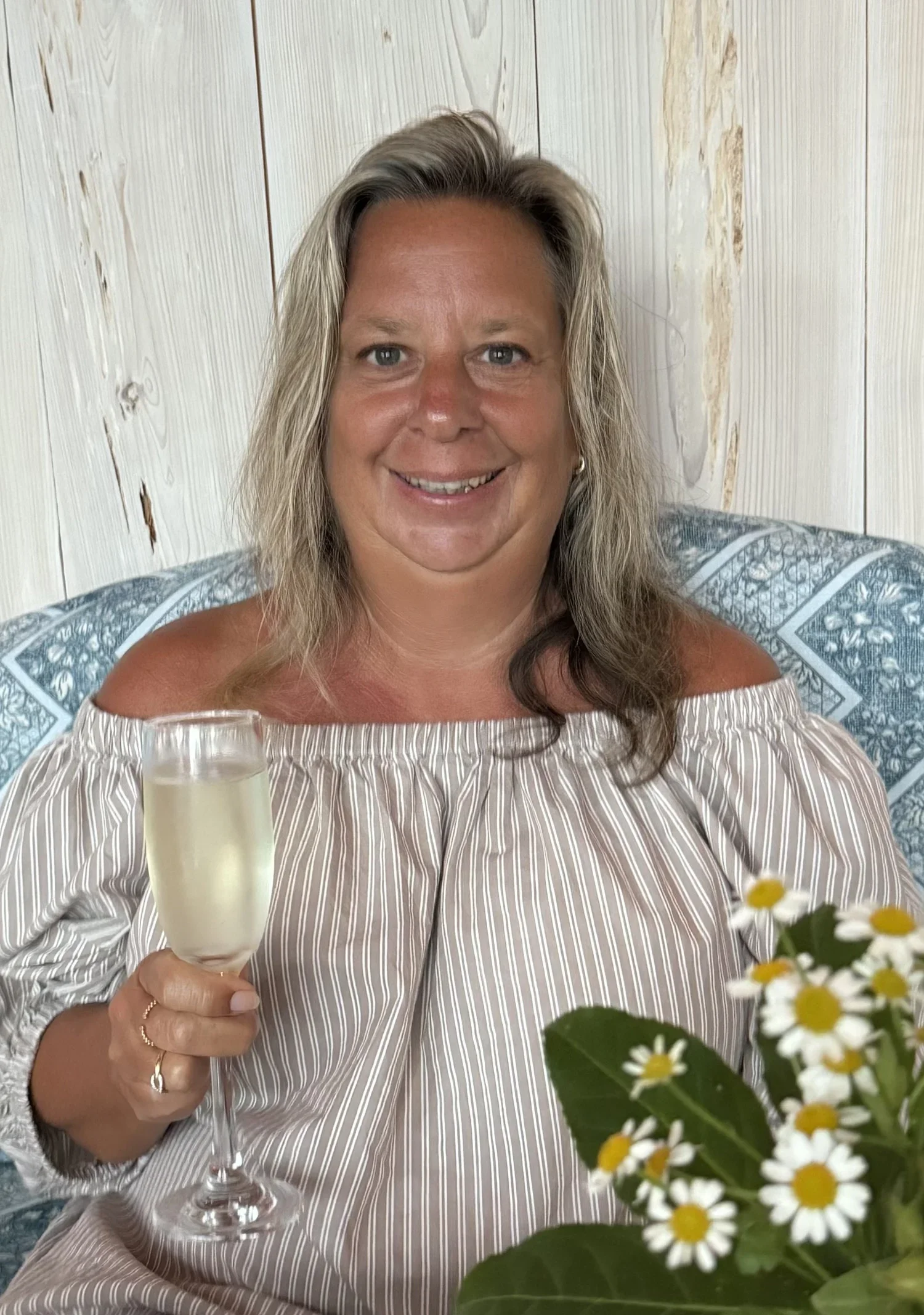 A woman with blonde hair smiling while holding a glass of champagne, sitting at a table with daisies in a vase, against a wooden wall background.