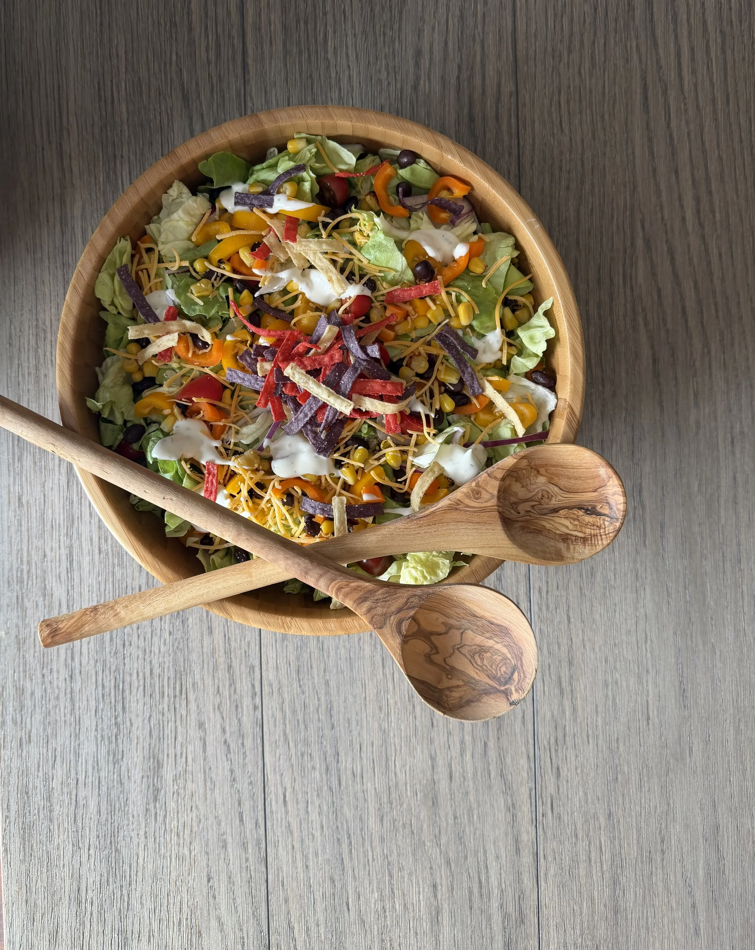 A wooden bowl of colorful salad with mixed greens, corn, shredded cheese, shredded beef, sliced bell peppers, cherry tomatoes, and dressing, on a gray wood surface with a wooden spoon.