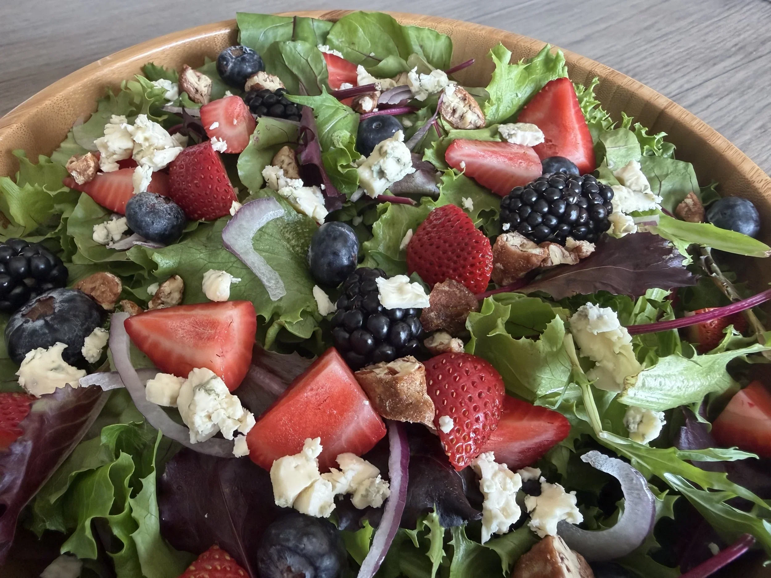 A salad in a wooden bowl containing mixed greens, strawberries, blueberries, blackberries, crumbled blue cheese, red onion slices, and walnuts.