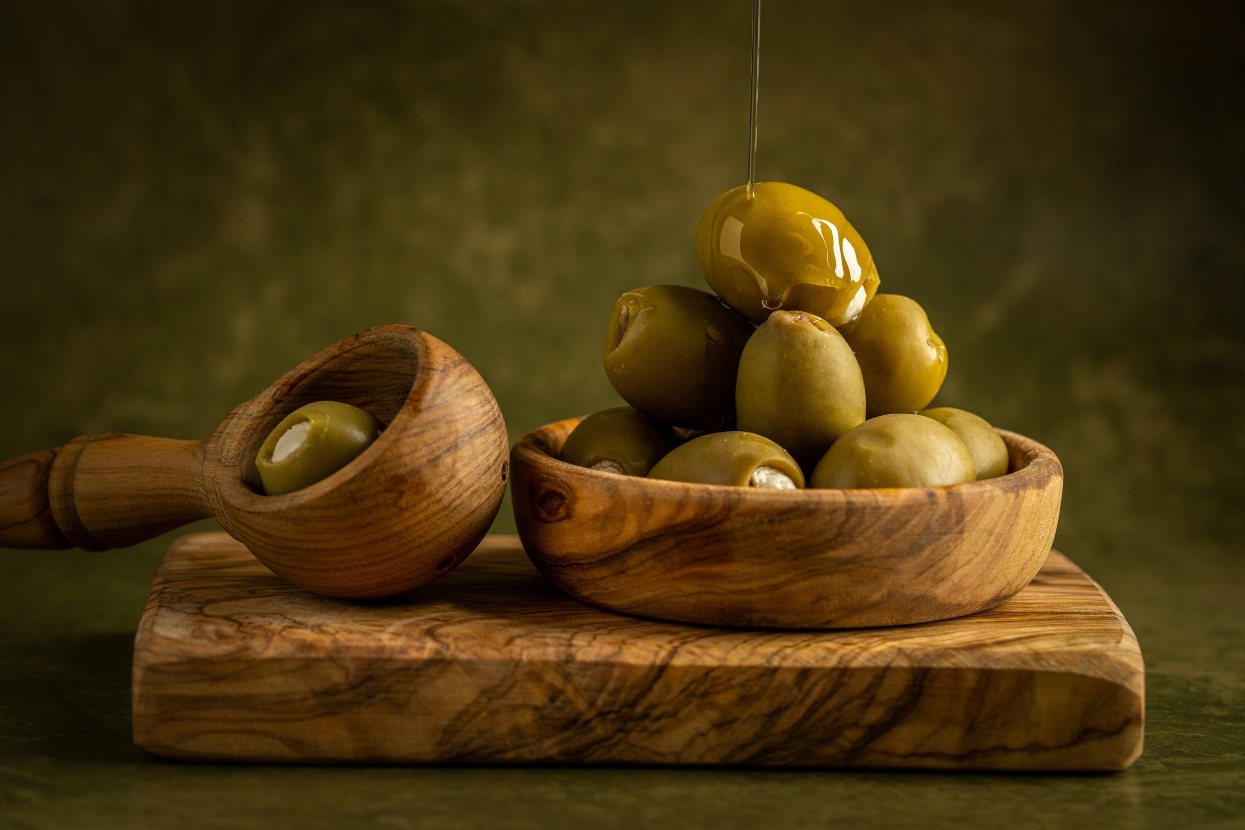 A wooden bowl filled with green olives, with one olive being drizzled with olive oil, all placed on a wooden surface with a dark green background.