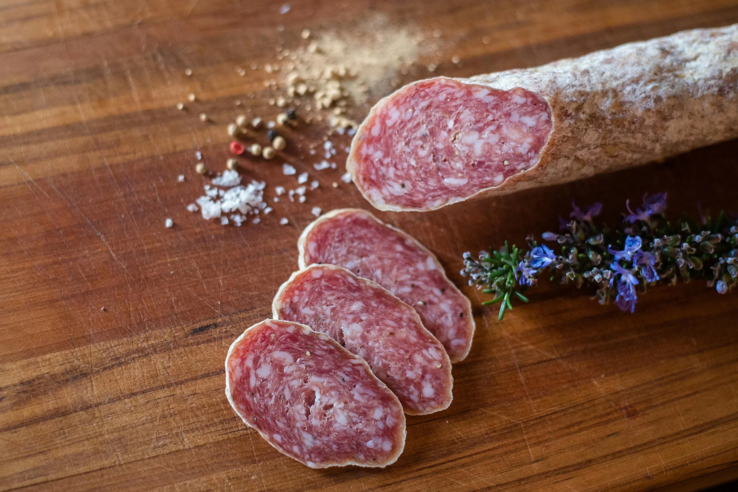 Slices of salami on a wooden cutting board with scattered salt, pepper, and herbs nearby.