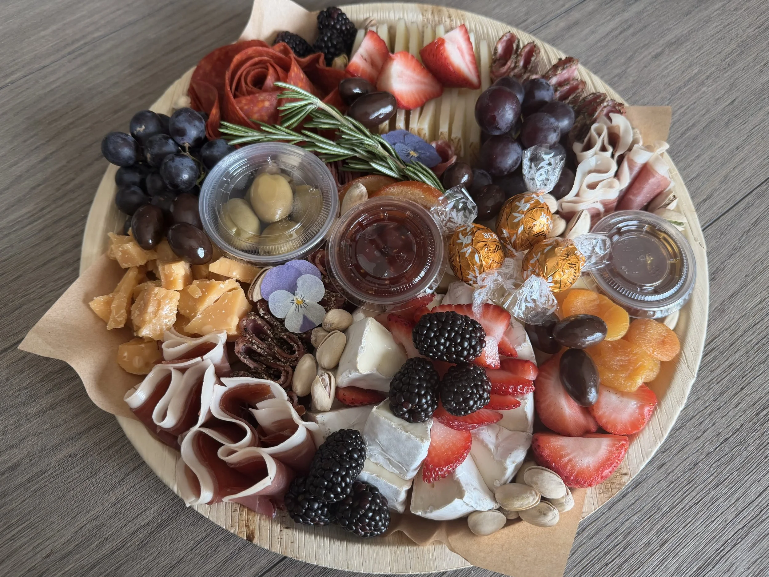 Round cheese platter with strawberries, blackberries, grapes, sliced cured meats, cheese slices, pistach nuts, honey or syrup in small containers, and garnished with sprigs of rosemary and edible flowers on a wooden board.