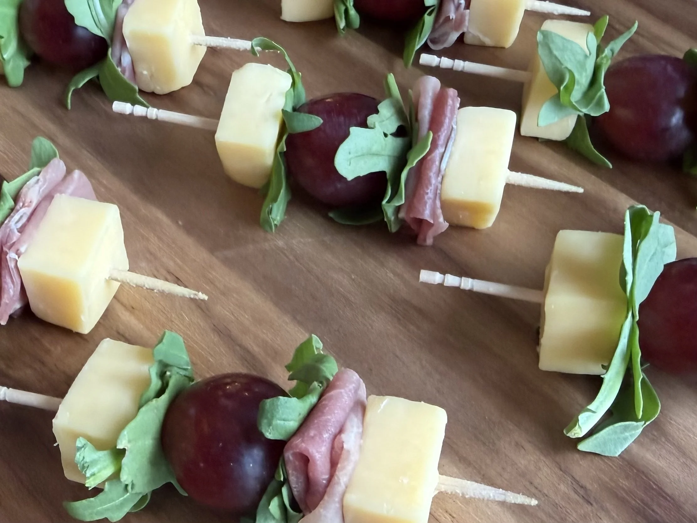 Close-up of several cheese and meat skewers topped with grapes, lettuce, and cheese blocks, arranged on a wooden surface.