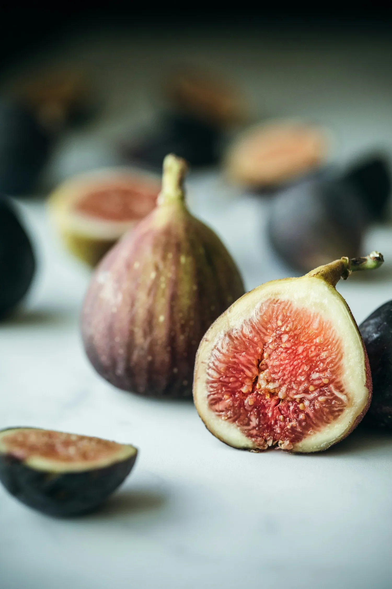 Close-up of cut figs on a white surface, showing the pink interior with seeds and greenish exterior, with whole figs in the background.