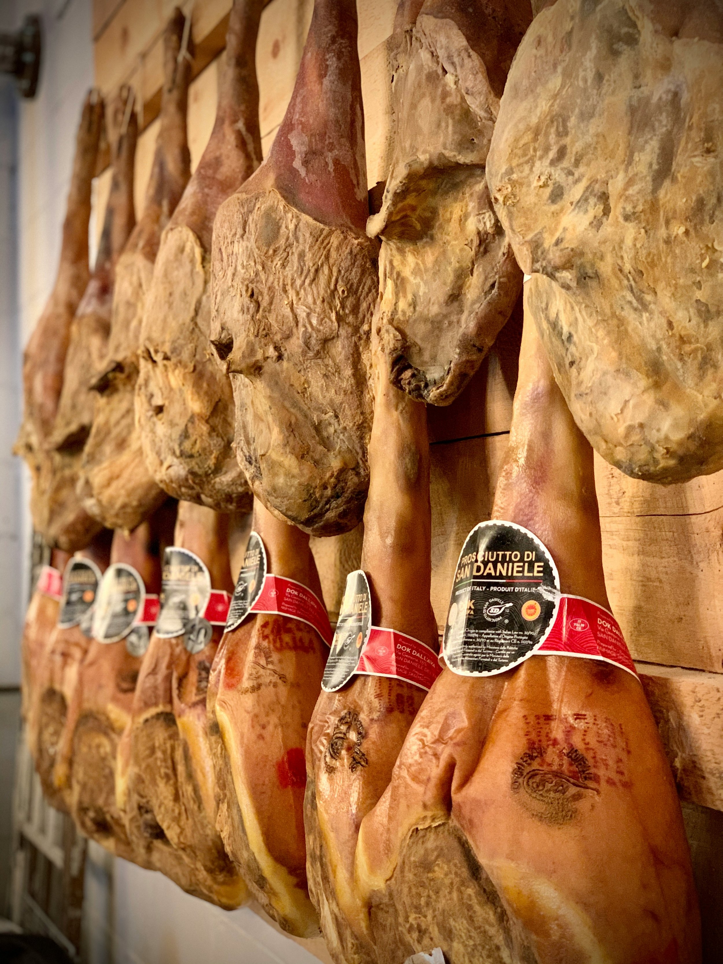 Close-up of cured ham legs hanging on a wooden wall, with labels attached, in a traditional Italian meat shop or deli.