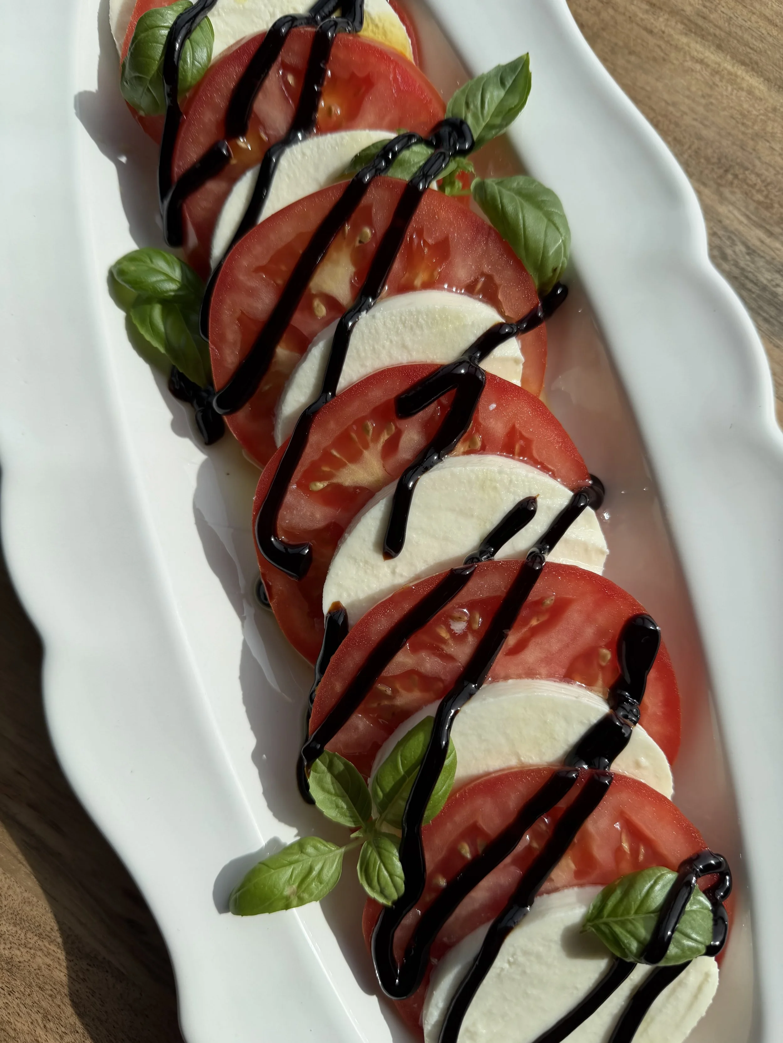 Caprese salad with sliced tomatoes, mozzarella, fresh basil leaves, and balsamic glaze on a white platter.